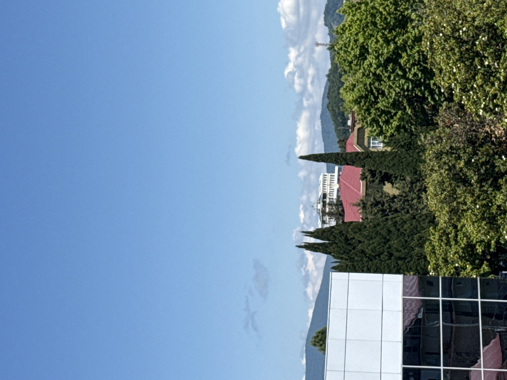 The image is a landscape view taken on a clear, sunny day with a bright blue sky and scattered clouds. The foreground features a modern building with a white facade and large glass windows, reflecting the surroundings. To the right of this building, several tall, dark green cypress trees stand prominently. Beyond the cypress trees, a red-roofed building is visible, along with more lush green trees and foliage. In the background, rolling green hills and mountains can be seen under the expansive sky. A glimpse of a large, white building with many windows and a domed structure is visible behind the cypress trees and red-roofed house. The scene appears to be a residential or commercial area in Sochi, Russia, with its characteristic blend of modern architecture and abundant greenery. No people are visible in the frame.