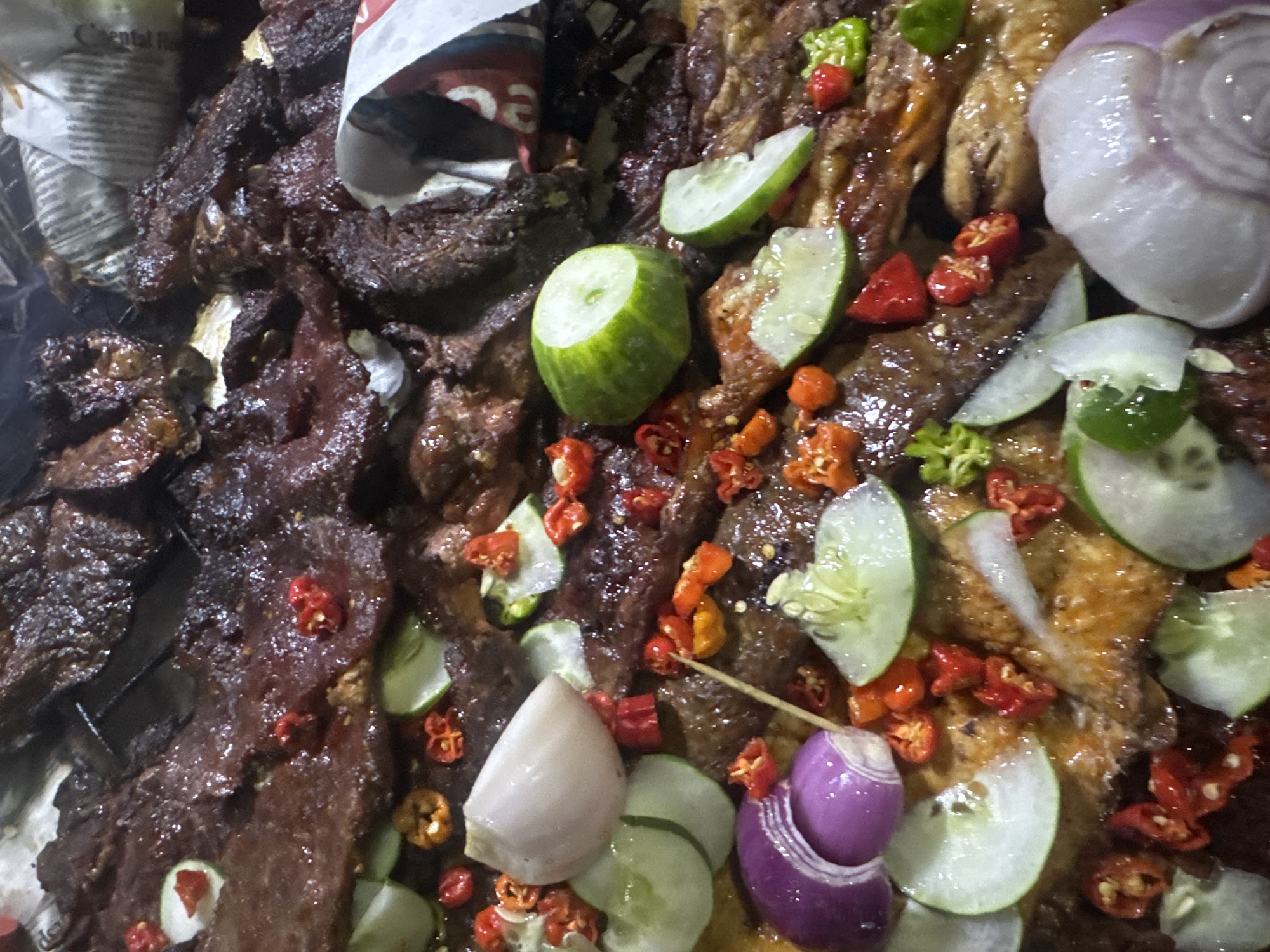 This is a close-up, overhead shot of a dish of grilled meat, likely beef, in Abuja, Nigeria. The meat is dark brown and glistening, suggesting it has been well-marinated and cooked. It is presented on a wire rack, possibly over coals, with some crumpled newspaper visible underneath. Scattered around the meat are slices of cucumber, thinly sliced red onion, and small, bright red chili peppers. There are also a few pieces of what appear to be raw garlic. The overall impression is of a flavorful and spicy street food dish. The lighting is dim, indicating it might be nighttime or an indoor setting.
