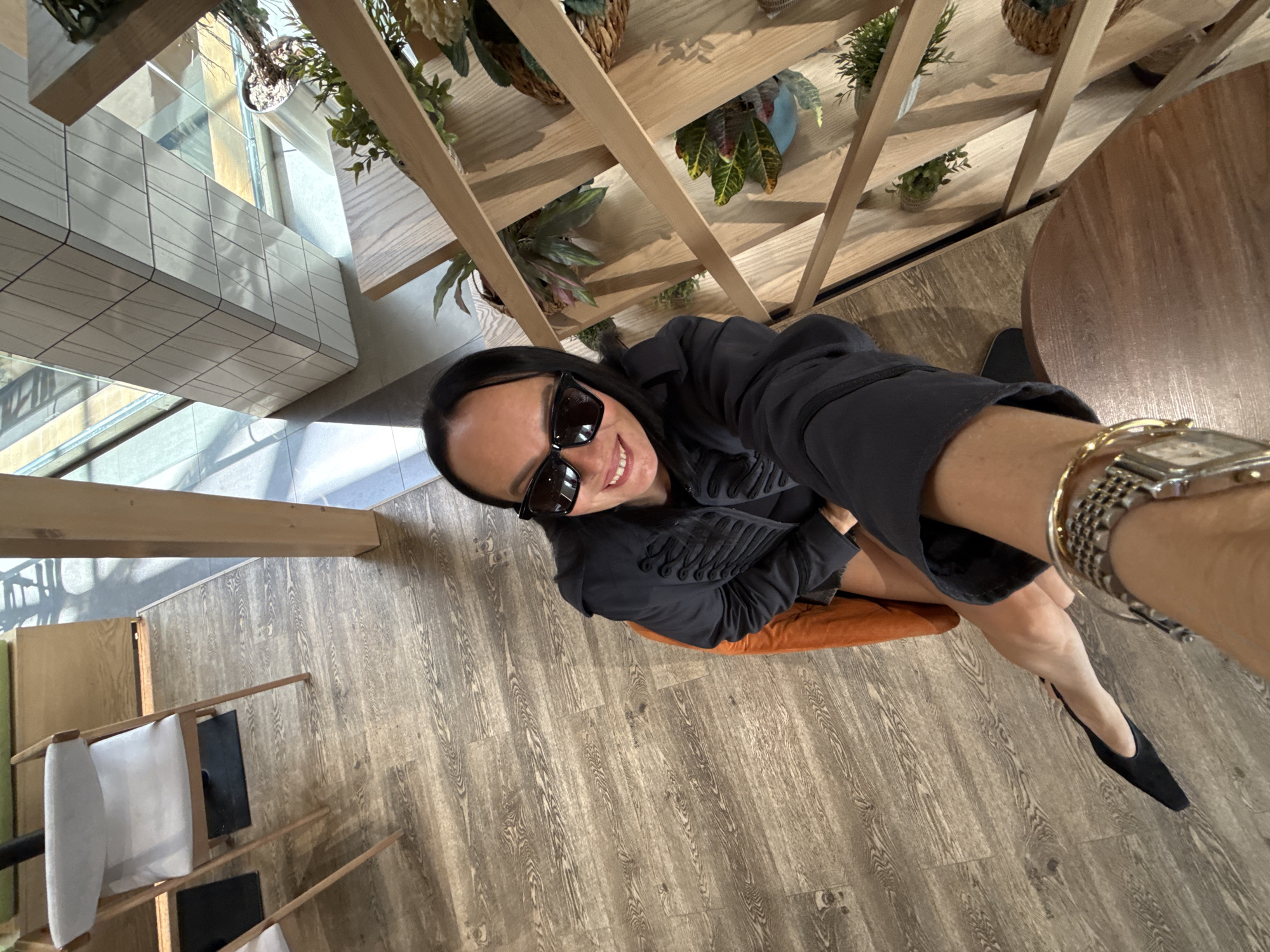 A woman wearing sunglasses and a black jacket is taking a selfie while sitting on an orange chair. She is in an indoor space with wooden floors and a wooden shelf unit filled with plants. A white chair and part of a table are visible in the background. The scene appears to be a cafe or a similar establishment.