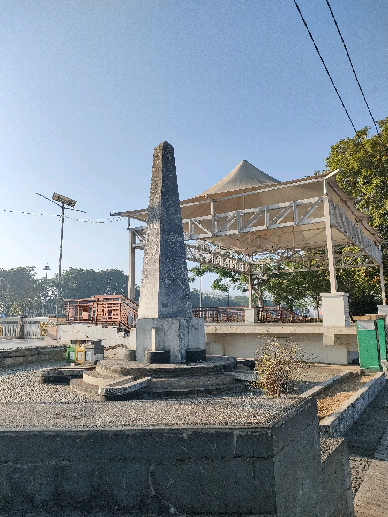 The image depicts the Siring Pierre Tendean Monument in Banjarmasin, Indonesia. The monument, an obelisk-like structure, stands on a tiered concrete base in the foreground. Behind it, a large, open-air pavilion with a conical roof supported by a metal frame is visible. The area appears to be a public space, possibly a waterfront promenade, given the presence of trees and a clear sky. The environment is bright and sunny, suggesting daytime. There are no people visible in the scene. The monument and pavilion are the primary man-made structures, while the surrounding environment includes paved surfaces, concrete barriers, and vegetation. The overall scene conveys a sense of openness and public accessibility.