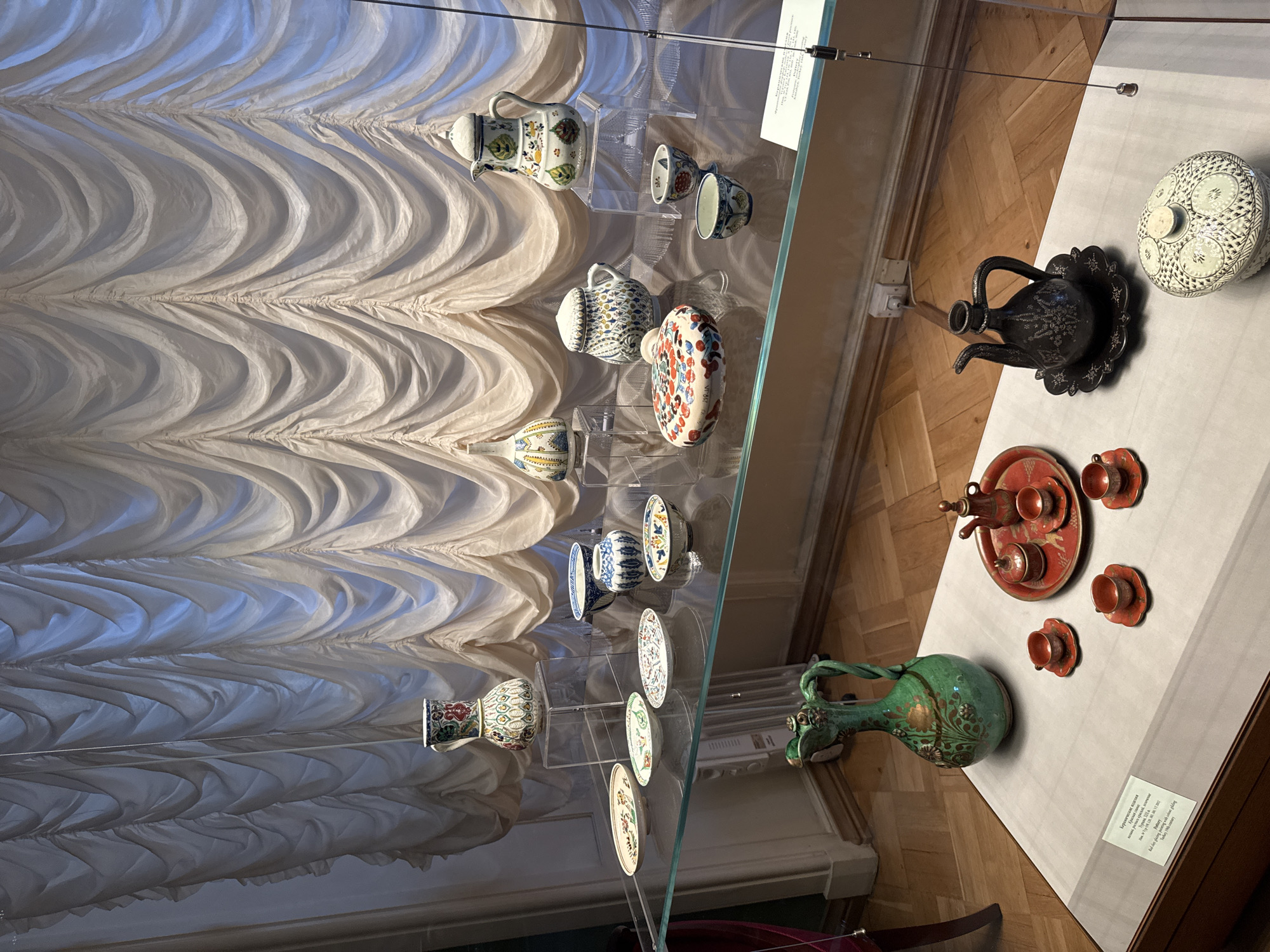 The image displays a collection of ceramics exhibited behind glass in a museum setting. The backdrop features draped, cream-colored curtains. On a glass shelf, various ceramic pieces are arranged, including bowls, plates, a teapot, and decorative vases, many adorned with intricate patterns and vibrant colors. Below this shelf, on a white surface, a green pitcher with a twisted handle sits prominently. To its right, a small red tray holds a teapot, sugar bowl, and several cups, forming a miniature tea set. Further to the right, a dark, ornate teapot with a flared base and a patterned lidded pot are displayed. A label is visible on the lower left, indicating the location is in Saint Petersburg, Russia, specifically the State Hermitage Museum, with some items identified as pottery from Turkey dating to the 16th-17th centuries. The floor is made of polished wood.