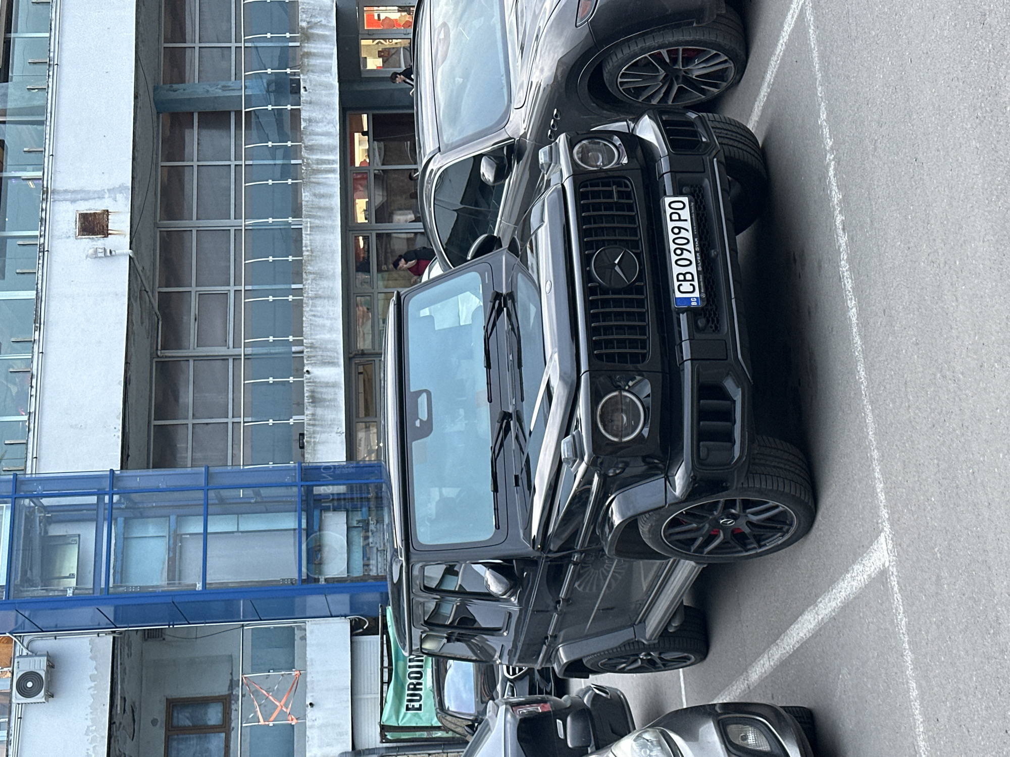 An outdoor, eye-level shot captures a black Mercedes-Benz G-Wagen parked in what appears to be a parking lot next to a multi-story building. The vehicle is positioned facing the viewer's left, with its front grille and headlights clearly visible. Behind the G-Wagen, a dark-colored SUV is partially parked, and another silver/grey vehicle is visible at the bottom of the frame.
The building on the left side of the image features a facade of concrete or stucco with numerous large glass windows, suggesting an office or commercial structure. A distinctive blue-framed glass-enclosed walkway connects different sections of the building higher up. Small details like an external air conditioning unit and a vent are visible on the building's exterior. Blurred figures of people can be discerned through the building's windows, indicating activity inside.
The parking lot surface is asphalt with white painted lines. The G-Wagen's license plate, "CB 0909 PO," is clearly visible, along with the country code "BG" and text "SILVER STAR www.silverstar.bg" below it, confirming the location as Bulgaria. The G-Wagen is fitted with large, dark alloy wheels featuring red brake calipers. Part of a green sign with "EUROIN" visible on it can be seen to the left of the G-Wagen's windshield. The lighting suggests an overcast day, providing soft, diffused light across the scene.