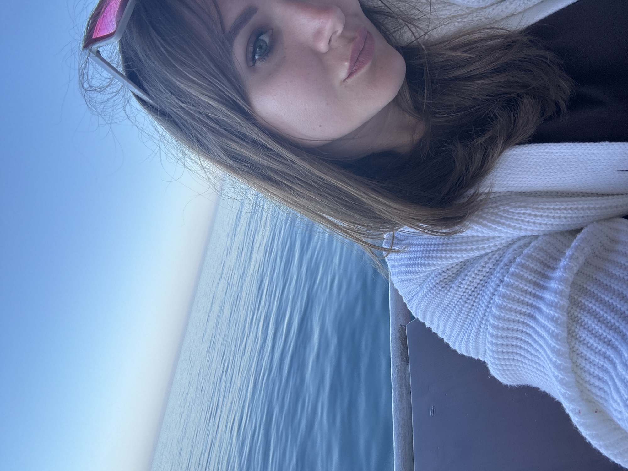 A woman is on what appears to be a pier or boardwalk next to a body of water, likely the Black Sea given the city mentioned. The woman is light-skinned, with long brown hair and pink-tinted sunglasses resting on her head. She is wearing a white knit sweater over a dark top. The water is blue and slightly choppy. The sky is a clear, pale blue. There are railings next to the pier. The photograph is taken at an angle, with the horizon line slanting diagonally across the frame. The photo is likely a selfie.