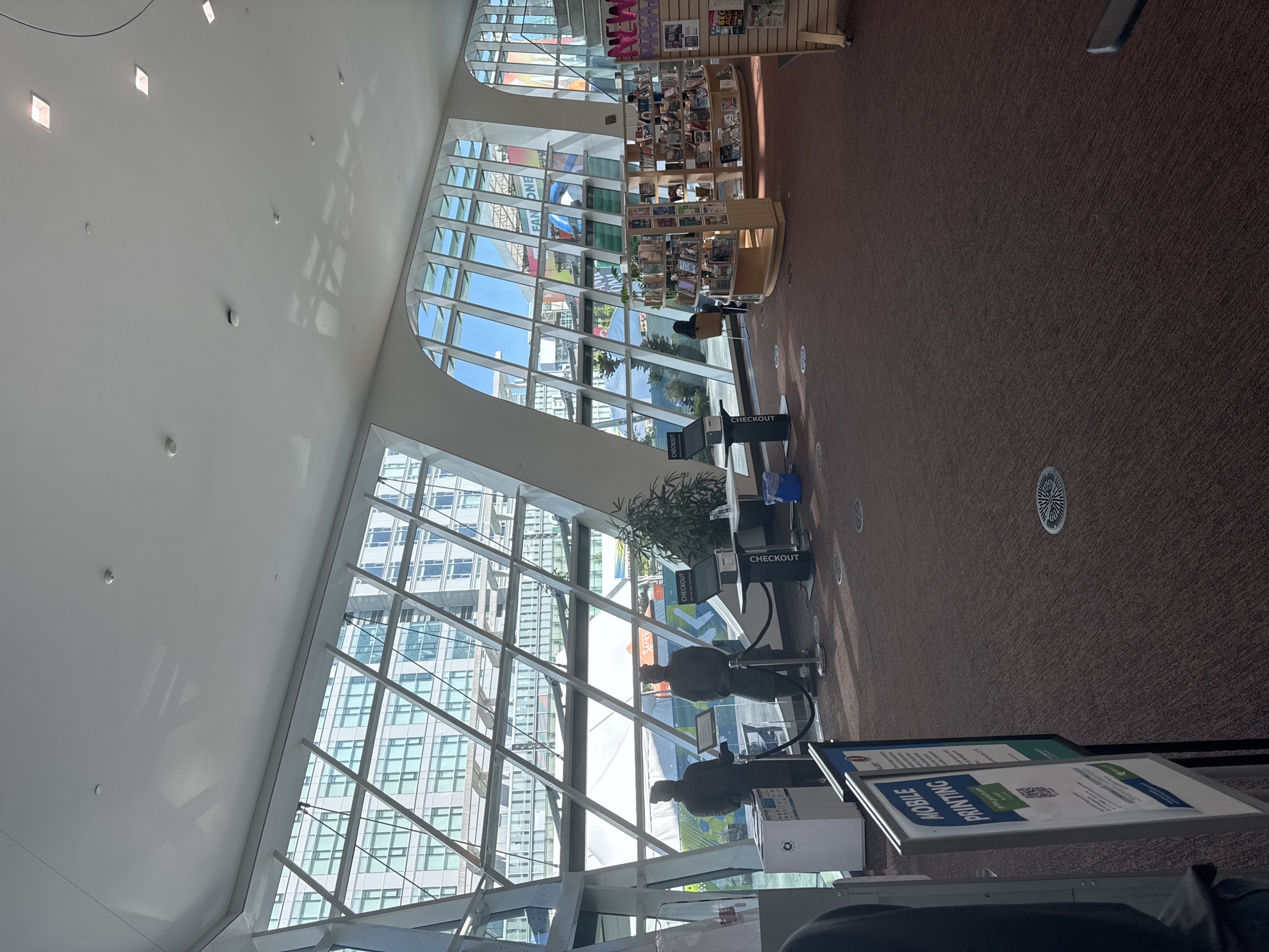 The image captures the interior of the Surrey City Hall, Canada, an architecturally modern building characterized by a large, angled glass facade. The space appears to be a public area, possibly a library or community hub, with a rich brown carpeted floor and a high, white ceiling equipped with numerous ceiling lights and circular ventilation grates.
In the foreground, to the left, a sign displays "MOBILE PRINTING" with a QR code and text, indicating a printing service. Two statues, resembling ancient warriors, stand near a checkout counter with a screen. Further into the space, a large potted plant sits near another checkout station.
The background reveals a row of bookshelves filled with books, and further to the right, a display rack showcases magazines. Beyond these elements, the expansive glass windows offer a view of an urban landscape with contemporary buildings. Sunlight streams through the glass, casting angular shadows across the interior.
One person is seated at a table in the mid-ground, facing away from the camera, engaged with a laptop. In the distance, another individual is visible near the bookshelves. Two figures, likely staff, stand near the warrior statues. The overall atmosphere is one of a bright, open, and functional public space.