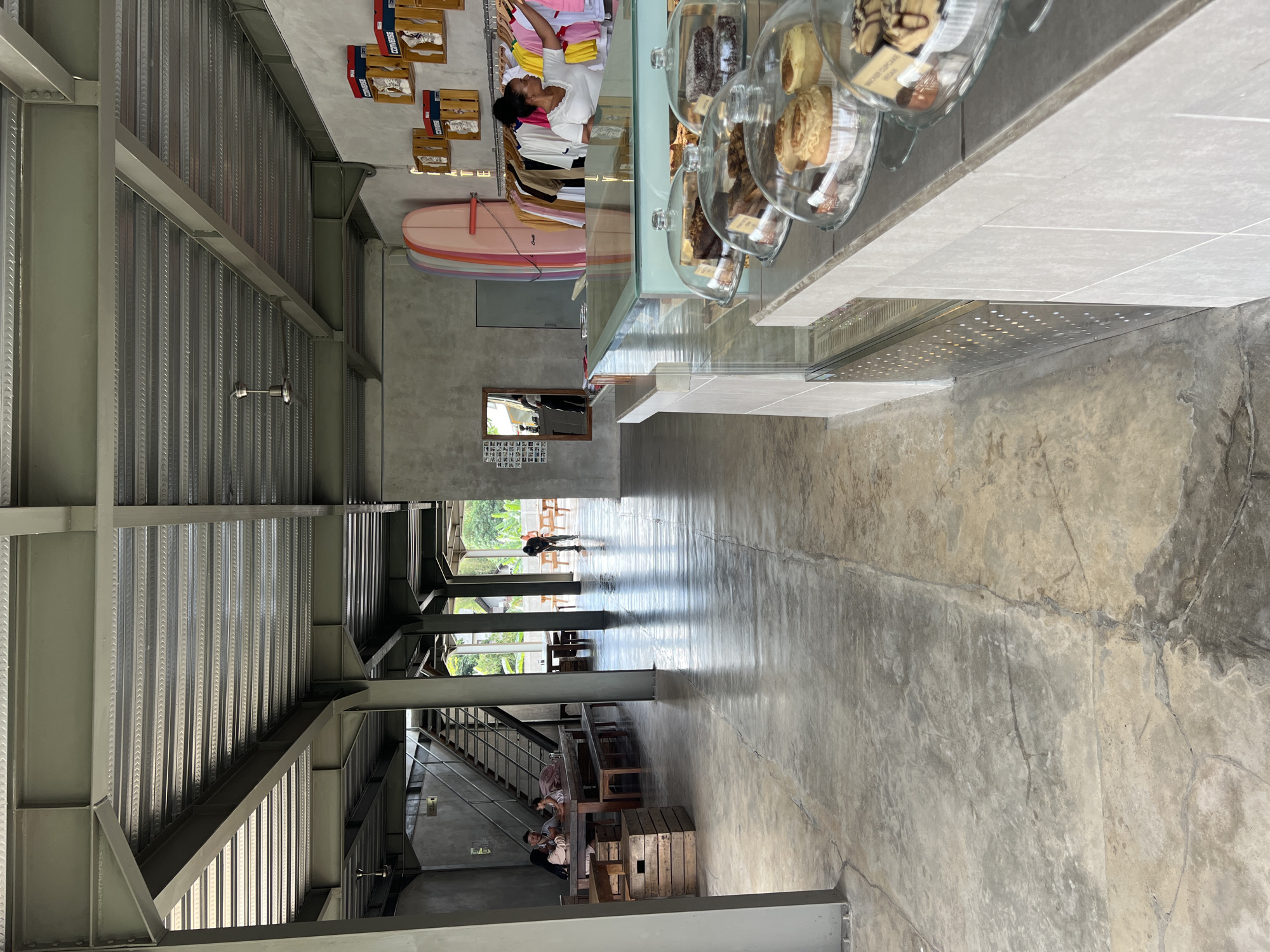 The image depicts the interior of a cafe in Mengwi, Indonesia. The space is characterized by exposed industrial elements, including a metal beam ceiling structure and concrete floors. In the foreground, a display counter features various pastries under glass domes, including cupcakes and brownies. Behind the counter, a woman is arranging clothing on a rack, with surfboards leaning against the wall and shelves displaying shoeboxes. In the background, a group of people are seated at tables, and another individual is standing near some tables. The cafe appears to be open-air or have large openings, as natural light illuminates the space and suggests an outdoor view.