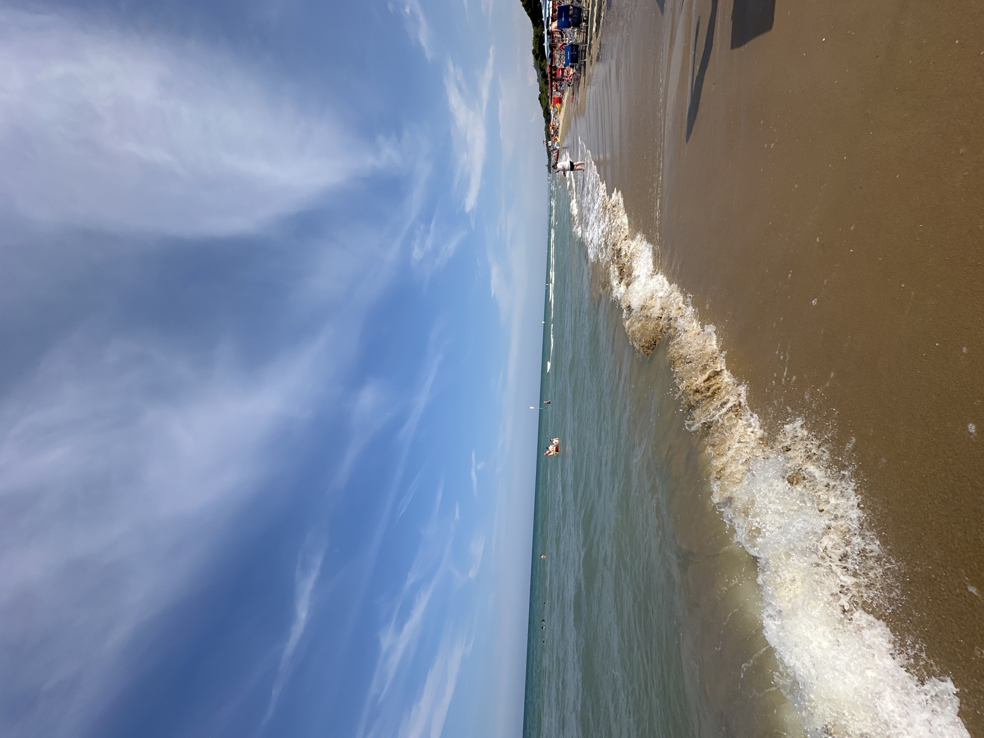 This is a wide-angle photograph of a beach scene in Pineto, Italy. The image captures a section of the Adriatic Sea meeting a sandy beach. The water is a greenish-blue with small waves breaking near the shore, creating white foam. Several people are visible in the water, some swimming and others wading.
On the sandy beach, there are rows of blue lounge chairs and umbrellas set up. A single person is standing near the water's edge, facing away from the camera. In the background, more beach umbrellas and a line of trees suggest the presence of a developed beachfront area. The sky is a bright blue with wispy white clouds. The overall impression is a sunny day at a popular beach resort.