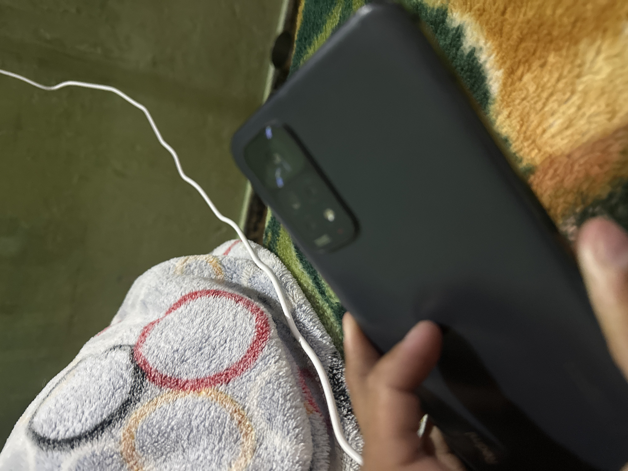 An interior scene depicts a hand holding a dark-colored smartphone, identified by a visible "Redmi" logo on its lower back, featuring a rectangular multi-lens camera module. A white charging cable extends into the frame from the upper right, partially obscured by the hand holding the phone. The phone rests on a textured, multi-colored rug with green, yellow, and potentially other hues. Behind the phone and cable, a light-grey fuzzy blanket or throw is visible, patterned with large, overlapping circles in red, black, and beige/gold. The background consists of a blurry, dark-green surface. The scene is situated in an interior setting, with the user specifying the location as the city of Juan Griego, Venezuela.