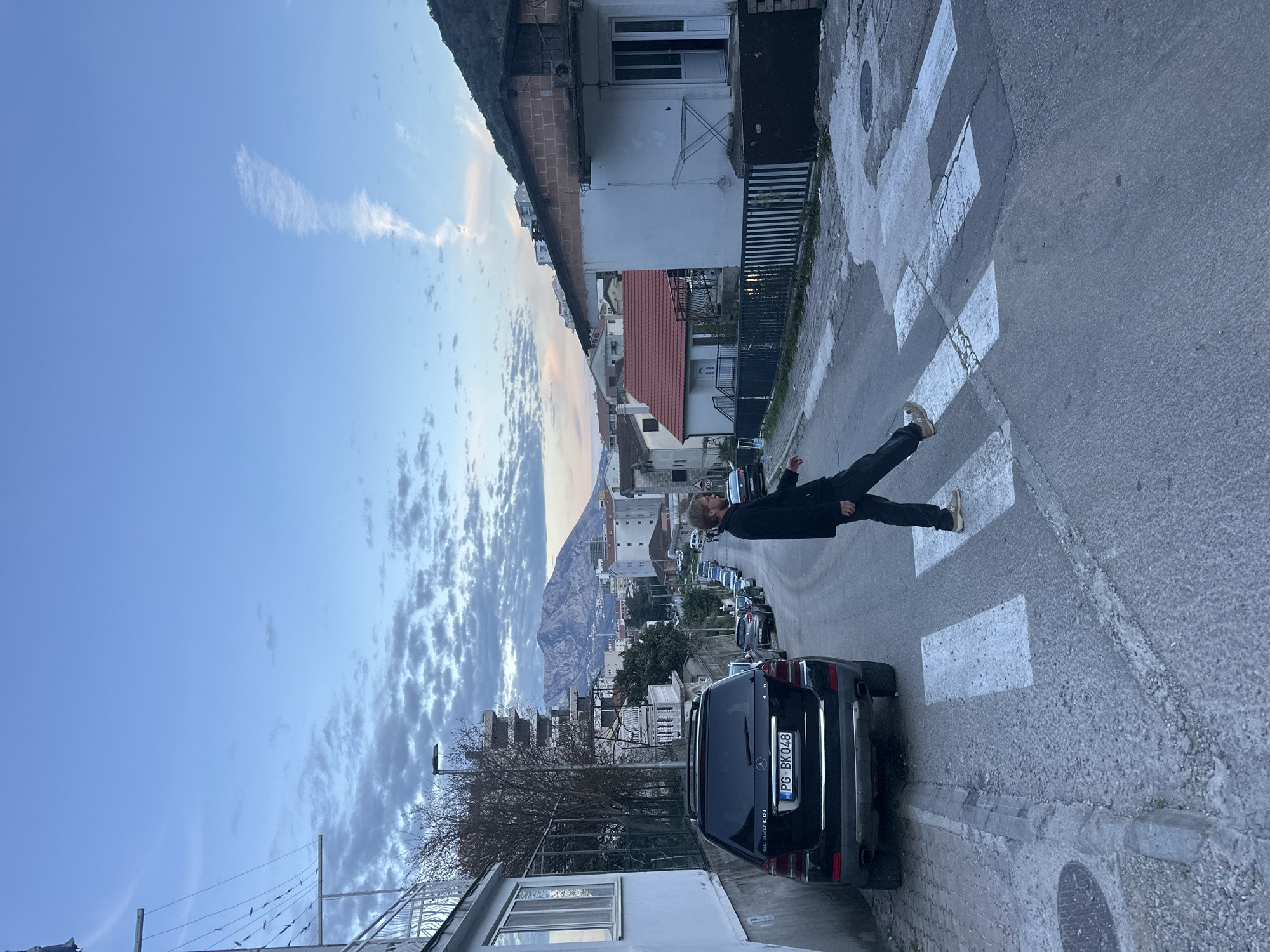 The image is a ground-level shot of a street in Budva, Montenegro. The street is asphalt, with a pedestrian crosswalk marked with white stripes. A black SUV is parked on the right side of the street, with the license plate "PG BK048" visible. A person, dressed in dark clothing, is positioned on the crosswalk, with one leg extended. The surrounding environment includes multi-story residential buildings on both sides of the street. In the background, there's a view of a mountain range under a sky with scattered clouds. Other parked vehicles can be seen further down the street.