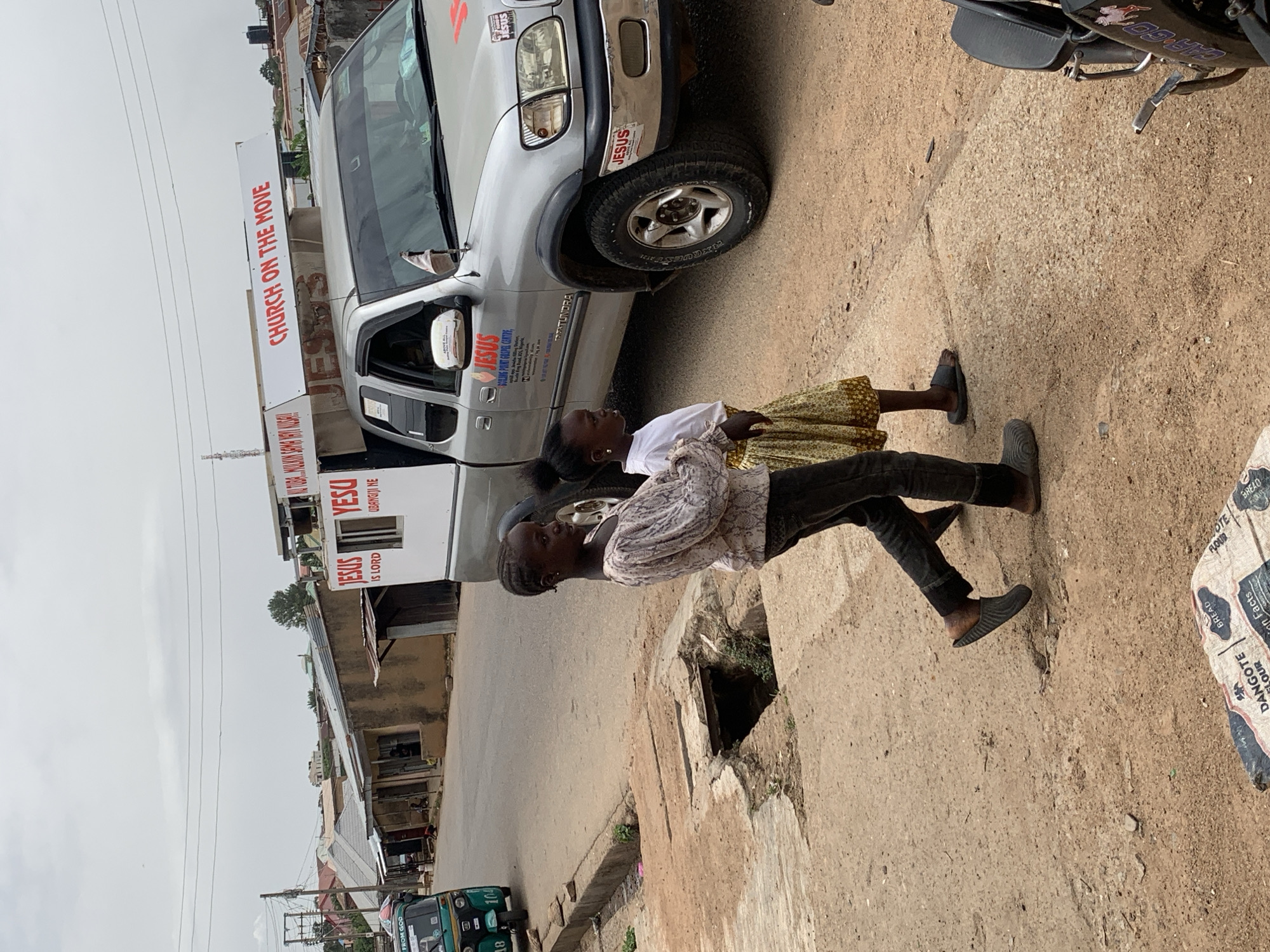 Here's a description of the image:
The image shows a street scene in Gura Top, Nigeria. Two children stand beside a drain on a dirt and concrete road near a silver Toyota Tundra pickup truck with Christian messages. The truck has several stickers on it, "Jesus is Lord," "Church on the Move," and "Jesus Point Gospel Centre." A small motorcycle is partially visible to the right. Opposite the pickup is a row of building fronts that appear to be storefronts. An auto rickshaw is at the edge of the street. A bag that says DANGOTE FLOUR is laying nearby.