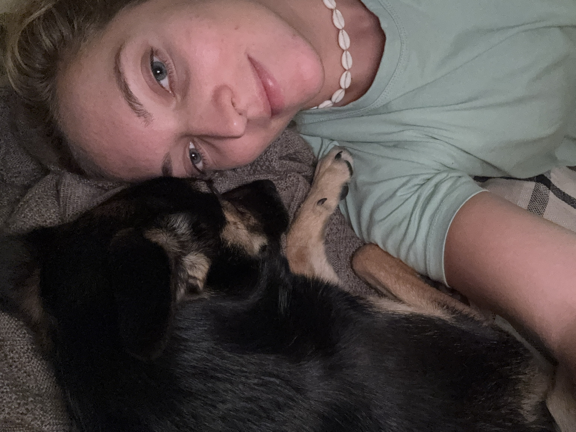 A woman with fair skin and light blue eyes is lying down with a small, black and tan dog. She is wearing a light green t-shirt and a white shell necklace. The dog is nestled close to her face. They are lying on a grey blanket or bedspread. The lighting is dim. There is no evidence to suggest this photo was taken in Minsk, Belarus.