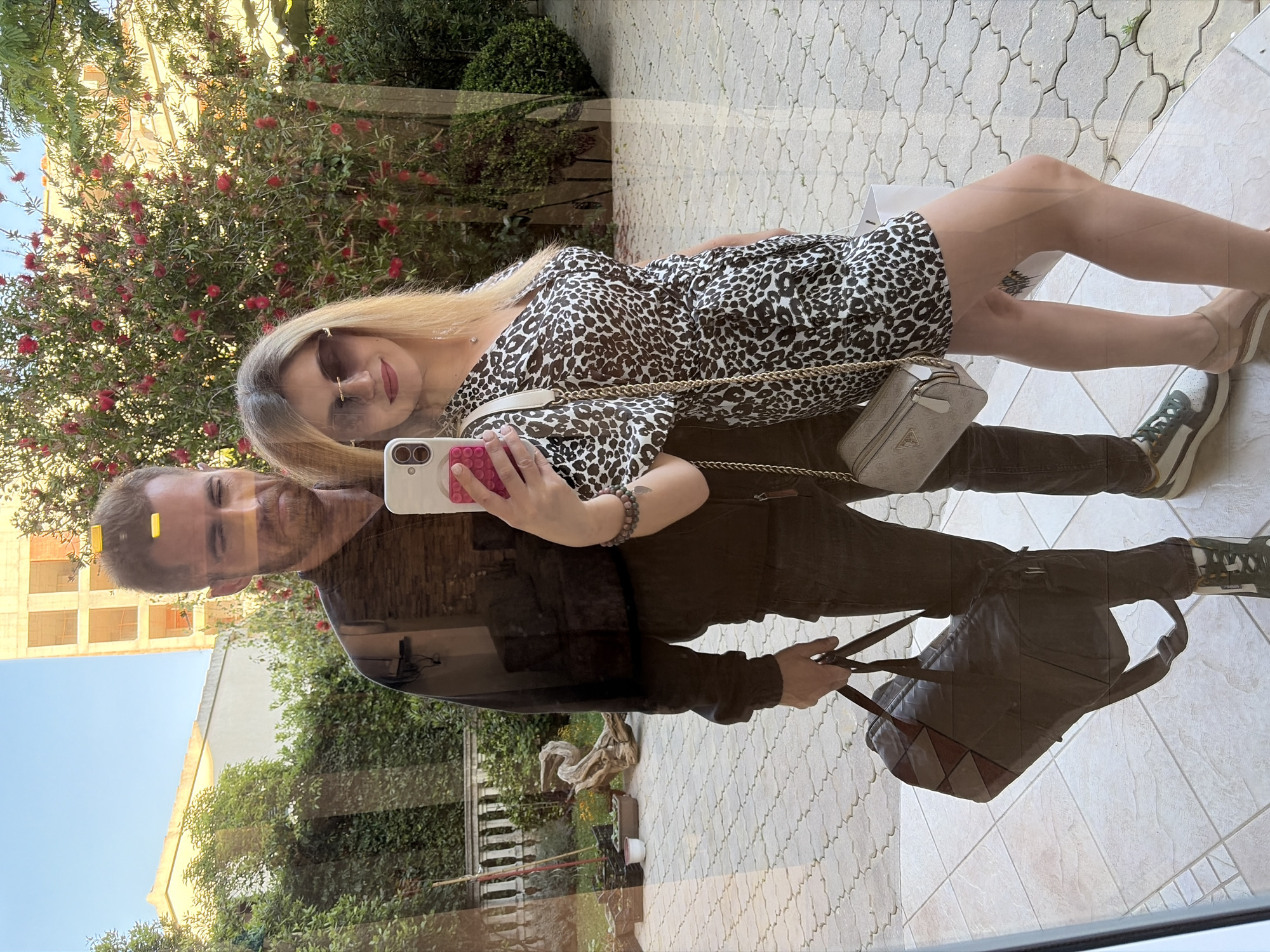 The image shows a reflection of a couple taking a selfie in front of a building in Budva, Montenegro. The woman is wearing sunglasses and a leopard-print dress with a white crossbody bag. She is holding a phone with a pink case to take the selfie. The man is wearing a dark-colored jacket and trousers and is carrying a large bag. The reflection shows trees, buildings, and patterned paving stones. The sky is visible at the top of the reflection.