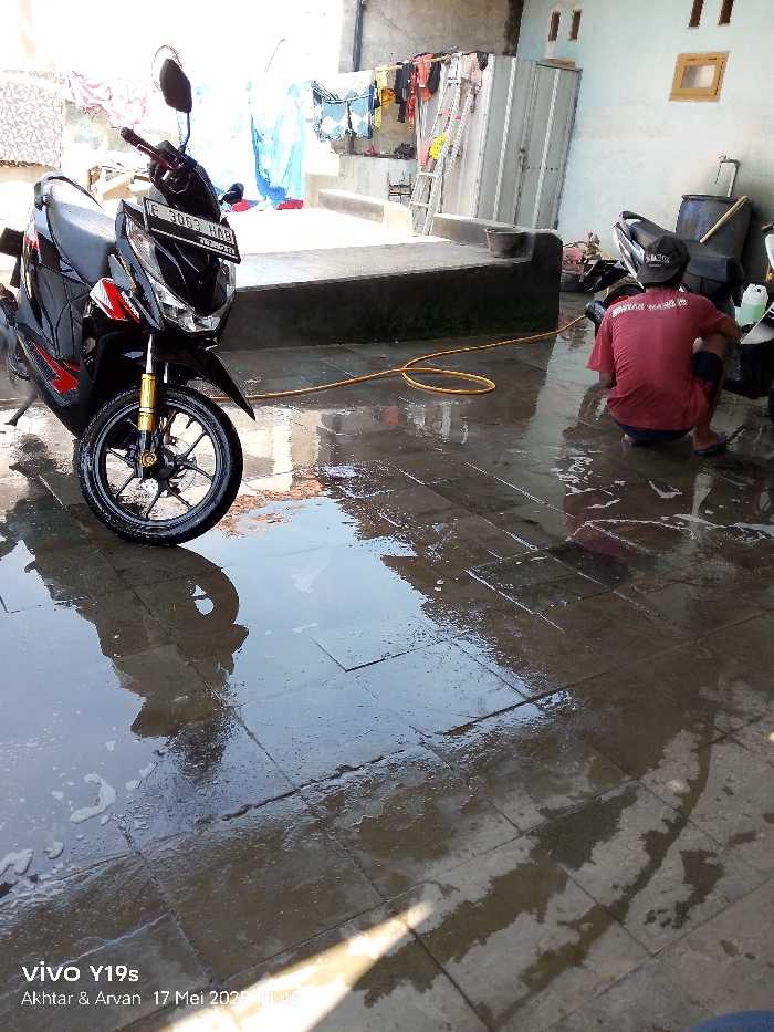 The image shows a wet patio in Gegesik, Indonesia, at the coordinates -6.533 latitude and 108.432 longitude. There is a black motorcycle parked on the left side of the frame. A person is sitting on the floor to the right of the motorcycle, cleaning the motorcycle. The floor is wet with soapy water and there is a yellow hose nearby. The surrounding environment is a patio with a wall and a metal gate on the left side and a building with a window on the right side. The patio appears to be constructed of stone pavers.