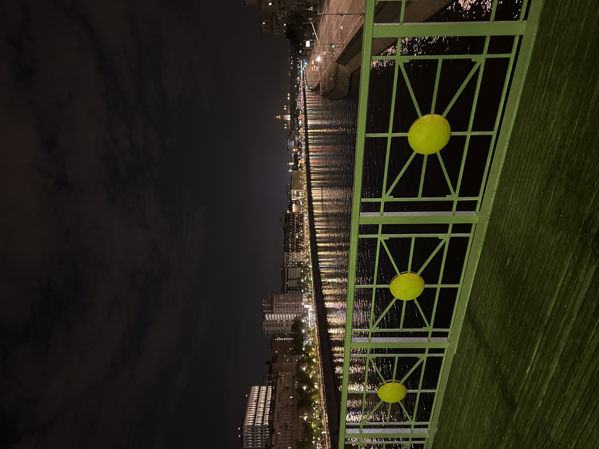 The image shows a nighttime view of a city from a bridge. The city lights reflect on the water below. A green metal railing with circular yellow accents is visible in the foreground. The sky is dark with some clouds. The location is Unknown, Unknown.