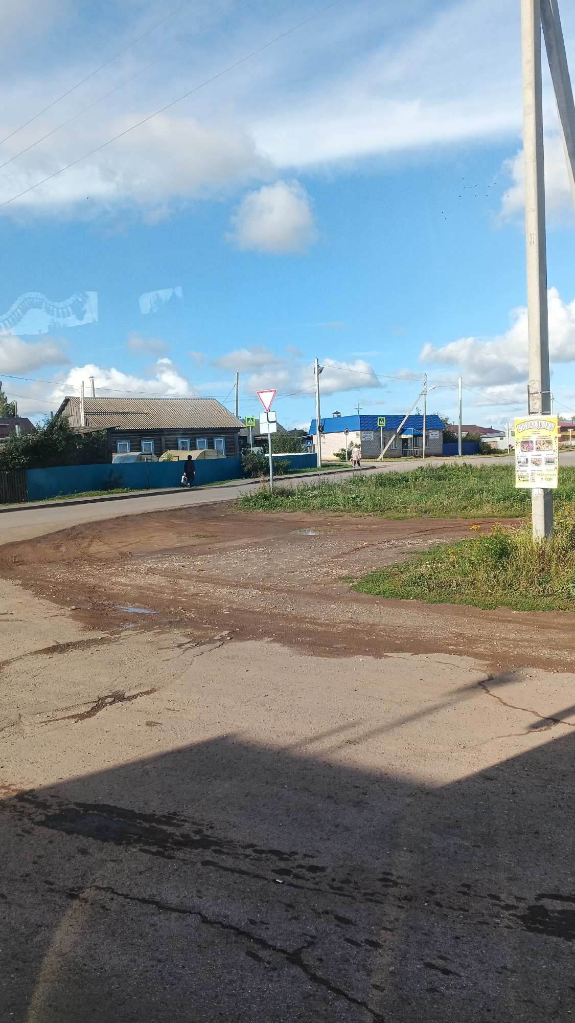 The image shows a street scene in the village of Kiyasovo, Russia. In the foreground, there is a dirt and asphalt road with puddles and tire tracks. A shadow of a structure is cast across the road. In the middle ground, there is a wooden house with a blue fence. Across the street, there is a blue building with a white awning and a signpost with traffic signs. A woman is walking in front of the blue building. Further in the background, there are more houses and a utility pole with a poster attached. The sky is blue with white clouds, and power lines are visible.