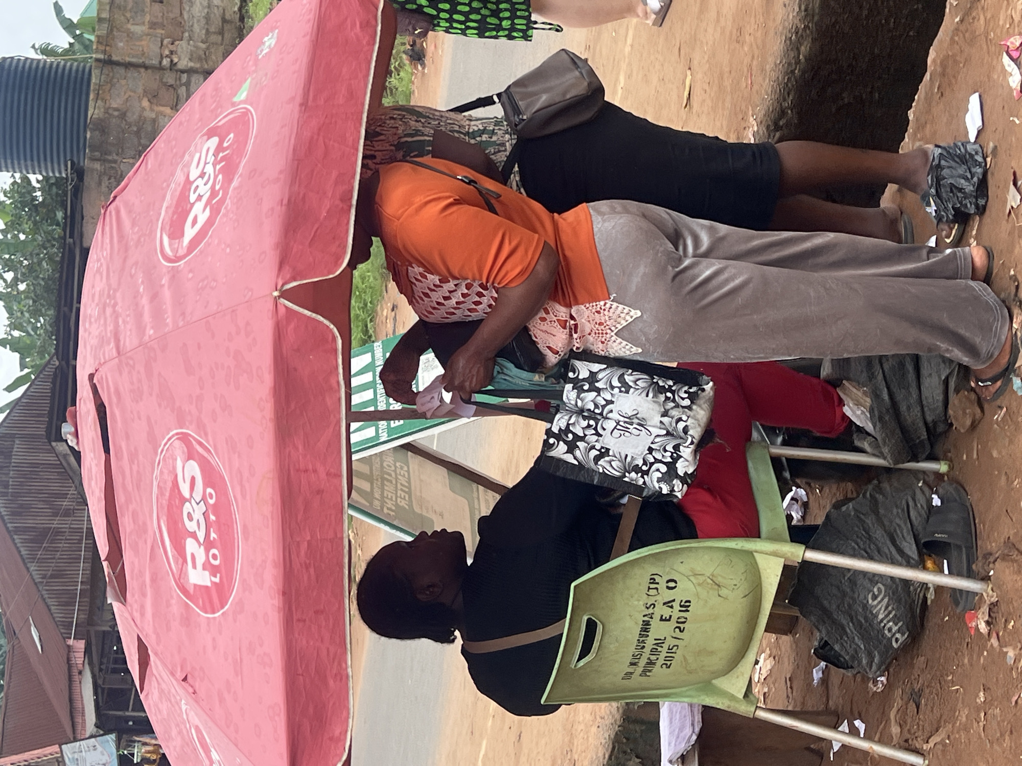 The image captures an outdoor scene in Nkpor, Nigeria, under a large red umbrella displaying the "R&S LOTTO" logo. Three individuals are visible. A woman in a black top sits on a green plastic chair, facing left, with a beige strap across her chest and a patterned tote bag on her lap. To her right, a woman in an orange top and grey pants stands, holding a bag and interacting with items. Behind her, another woman in a patterned top and dark pants stands, with a dark purse slung over her shoulder. The ground is dirt and littered with debris, including plastic bags and discarded paper. In the background, buildings with corrugated iron roofs and a water tank are visible. A sign with text, some of which appears to be in reverse, is propped up behind the seated woman.