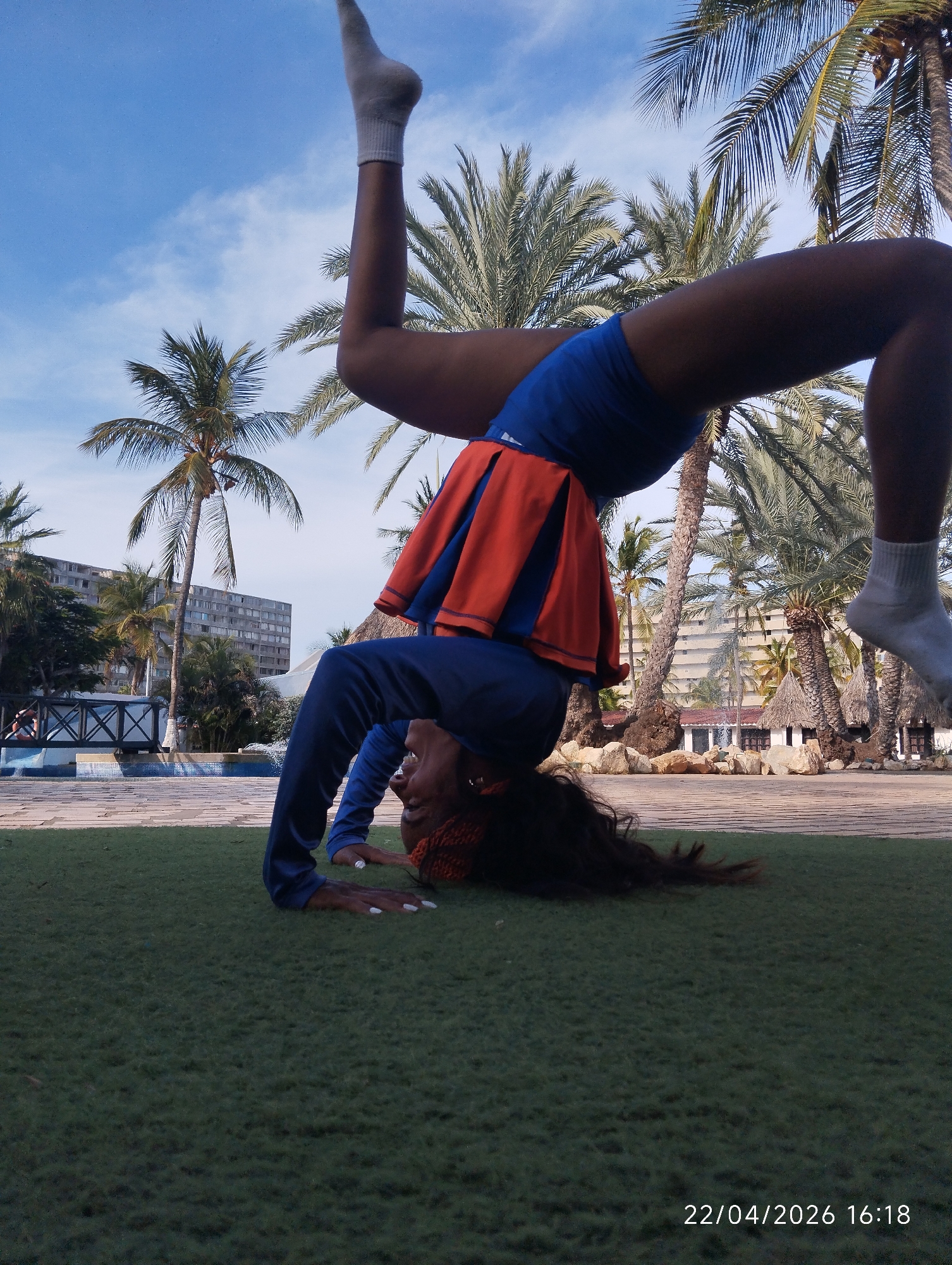 An outdoor, low-angle shot captures a person performing an acrobatic backbend or bridge pose on a patch of green, possibly artificial, grass. The individual, who appears to be a woman with a dark skin tone, is wearing a blue long-sleeved top, blue shorts, and an orange pleated skirt-like overlay. White socks are visible on both feet. Her hands are placed on the ground for support, with white-painted fingernails visible. Her long dark hair is fanned out on the grass, and her face is visible, looking upwards with an open-mouthed expression that suggests exertion or joy. Her body forms an arch, with her head resting on the ground, and one leg is bent at the knee while the other is extended vertically into the air.
The setting is tropical, characterized by numerous tall palm trees filling the background and reaching into the bright blue sky, which has scattered white clouds. Beyond the paved area adjacent to the grass, various structures are visible. These include multi-story buildings, possibly hotels or apartments, and smaller white structures with thatched roofs, indicative of resort-style architecture. A blue-tiled pool area and a decorative wooden bridge-like feature are also in the mid-ground. The lighting suggests it is daytime, confirmed by the timestamp.
In the bottom right corner of the image, the text "22/04/2026 16:18" is visible.