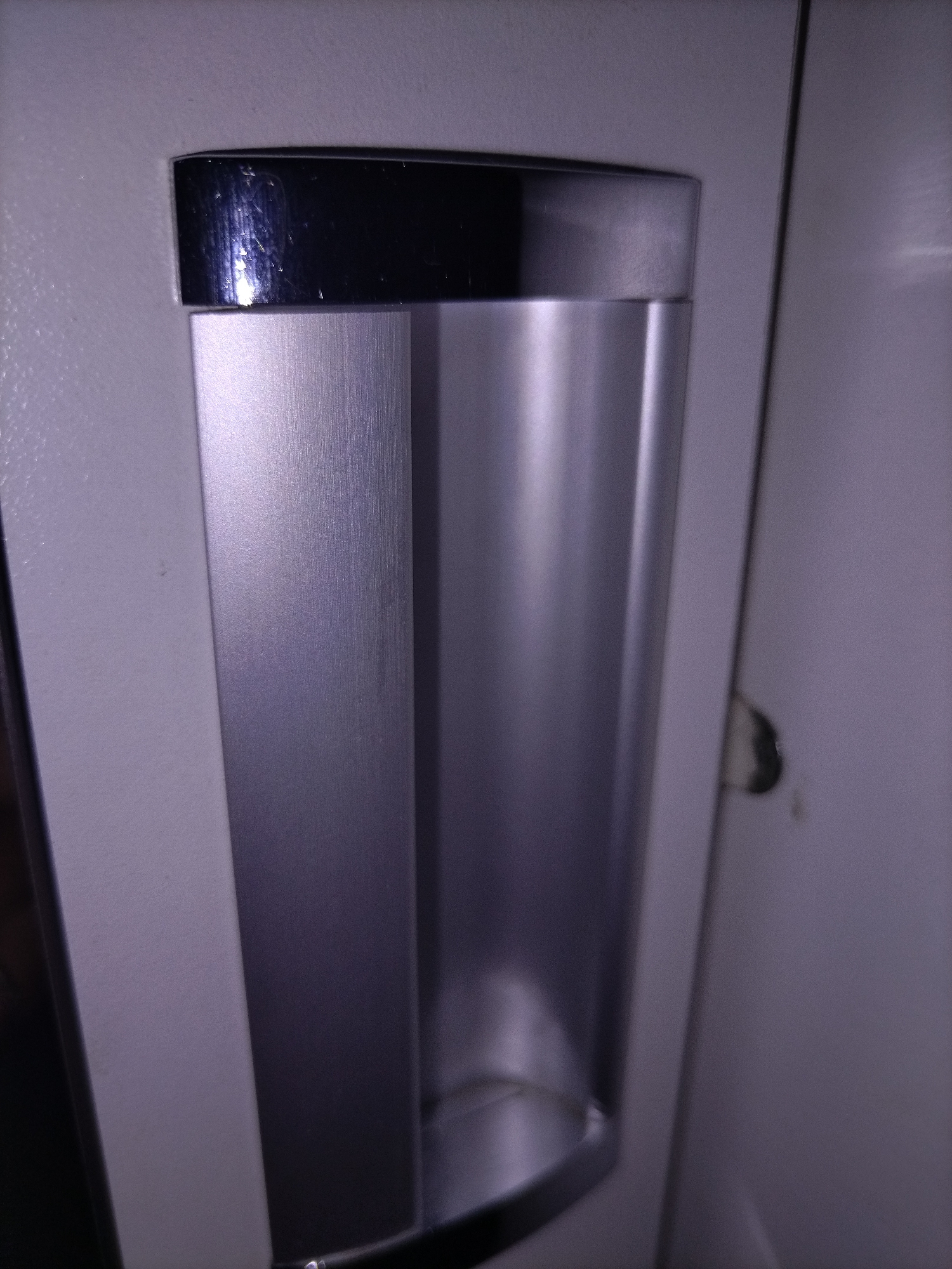 A close-up, slightly angled, eye-level shot shows a metallic cup holder attached to a light-colored vertical surface, likely a wall or cabinet. The cup holder has a sleek, modern design with a brushed aluminum finish, featuring a darker, reflective metallic rim at the top. The holder is elongated and appears to be hollow, with a curved bottom. The surrounding surface is textured and appears to be a light gray or off-white. To the right of the cup holder, there is a thin vertical seam, suggesting it's part of a larger panel or door. A small, dark, oval-shaped object is visible on the surface to the right of the seam, possibly a scuff mark or a small stain. The lighting in the image is dim, creating subtle shadows and highlights on the metallic surfaces. The background is out of focus, with muted colors.