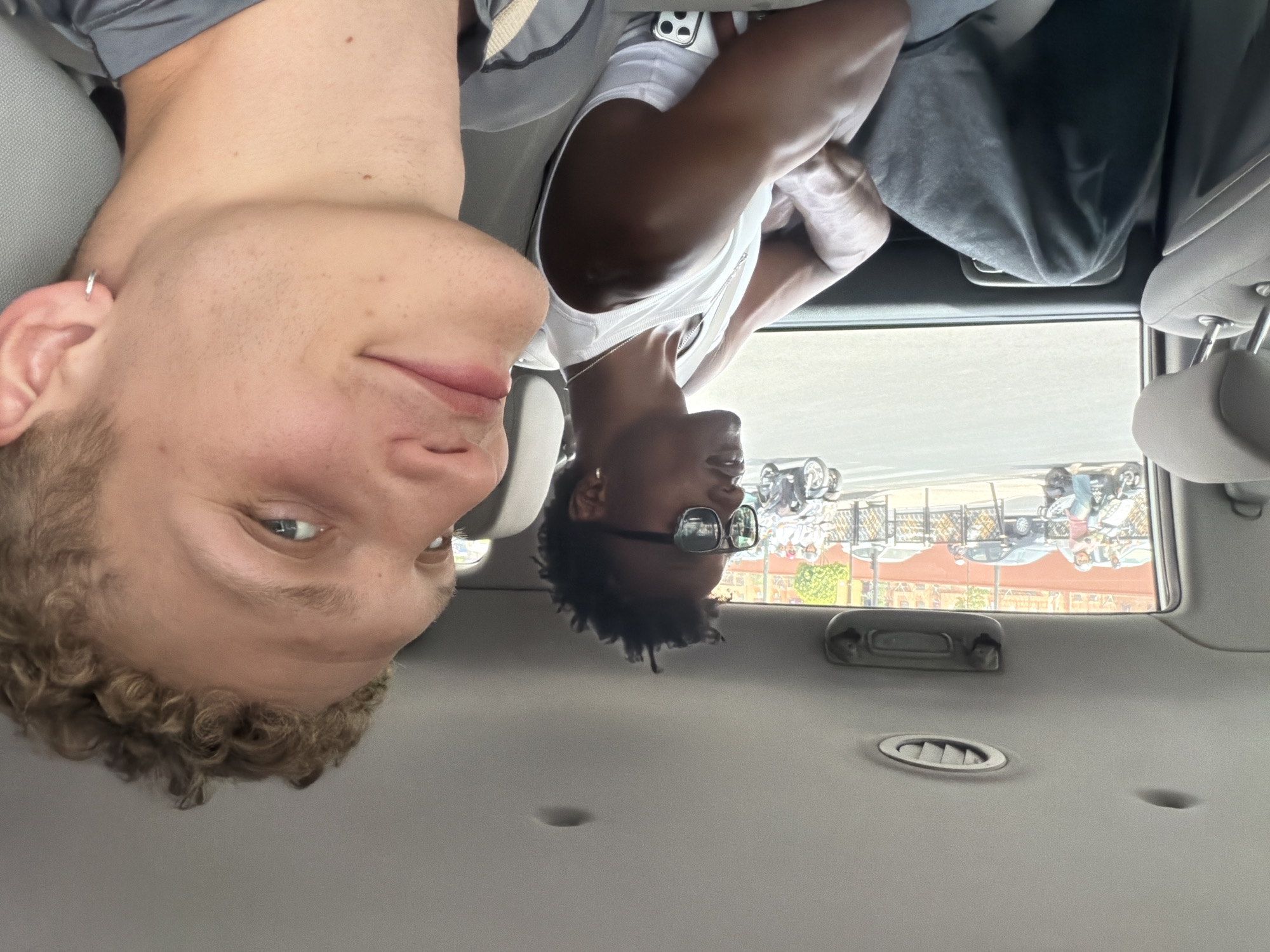 Two young men are seated in the back of a car, looking out the windows. The man on the right, in the foreground, has light skin, curly blonde hair, and a silver hoop earring. He is wearing a grey shirt. The man on the left has dark skin, short dreadlocks, and is wearing sunglasses and a white tank top with a silver chain. He has a silver hoop earring. Outside the car window, a street scene in Marrakech, Morocco, is visible with several motorbikes, cars, and buildings.