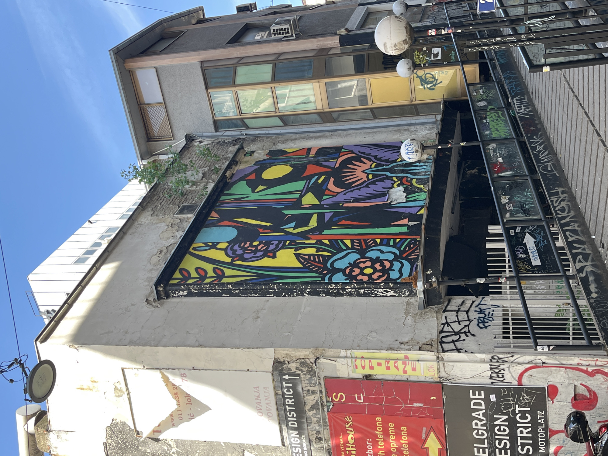 This photograph captures a narrow street scene in Belgrade, Serbia, showcasing a mixture of urban art, aging architecture, and street elements. The focal point is a large, colorful mural painted on a wall, exhibiting vibrant abstract floral and avian motifs. The mural is framed by a weathered building facade, which shows signs of deterioration with peeling paint and exposed brickwork. Below the mural, there's a collage of signage and graffiti, including "Belgrade Design District" signs.
To the right of the mural, a metal structure, possibly a ladder or scaffolding, is heavily marked with graffiti and stickers. Above, three white globes, likely light fixtures, are installed. Various advertisements and posters are plastered on the lower walls. The composition of the image suggests a tight urban space, with buildings densely packed together. The presence of graffiti and street art gives the area a bohemian, artistic vibe.