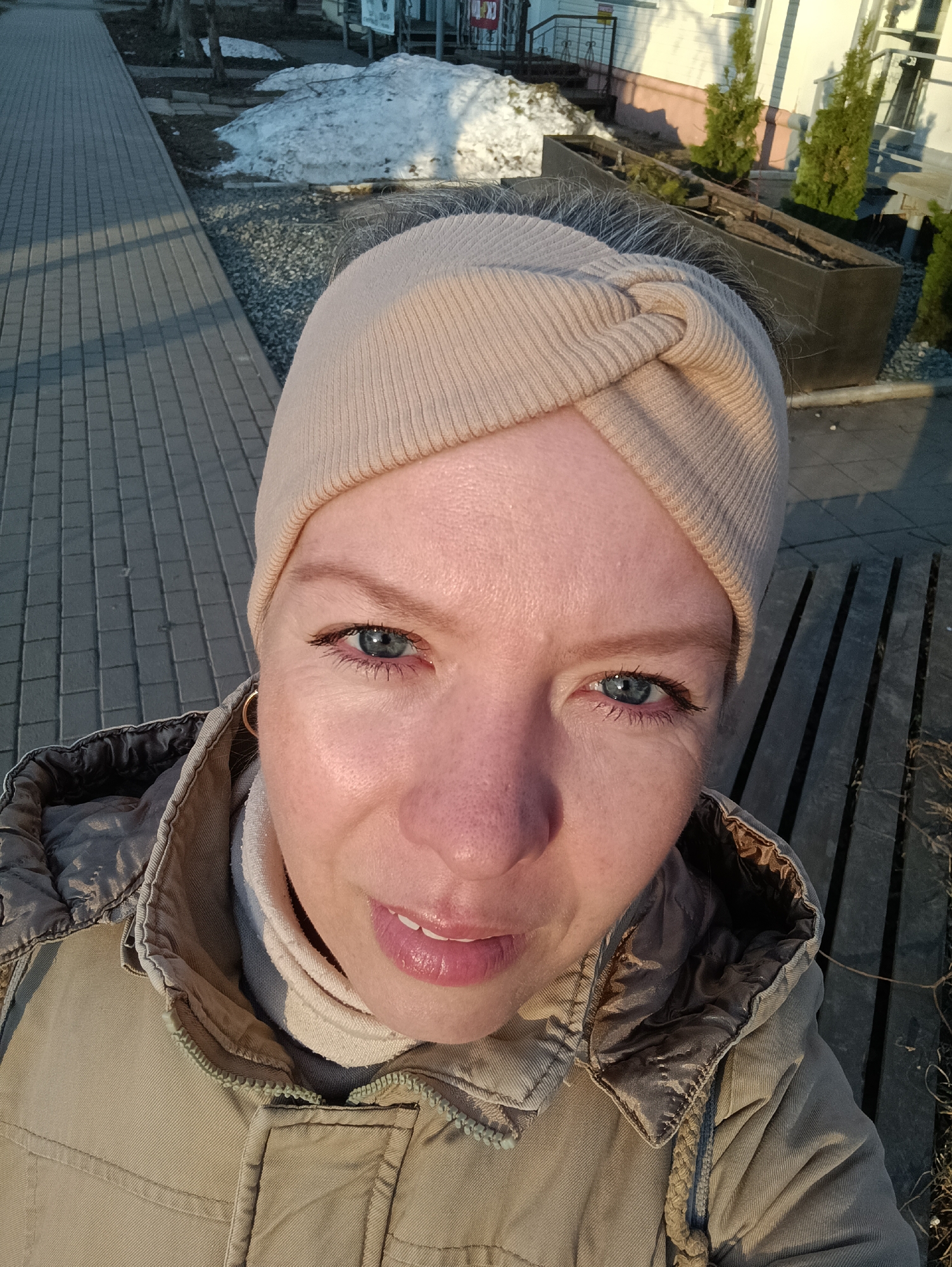 A high-angle selfie captures a woman outdoors in an urban or suburban setting, likely during late winter or early spring in Chaikovsky, Russia. She is the central subject, looking directly at the camera with clear blue eyes and a slight, pleasant smile. Her face is well-lit by warm, low-angle sunlight, suggesting either early morning or late afternoon. She has light skin, with some fine lines visible around her eyes.
The woman is wearing a light beige knit headband with a distinctive twisted knot design on her forehead, covering her hair, though some lighter strands are visible at her temples. She also wears a light-colored turtleneck and a light olive-green winter jacket with a quilted collar and a visible zipper. A small gold hoop earring is visible in her left ear.
The background reveals a paved sidewalk made of grey rectangular pavers on her left, running alongside a narrow strip of gravel. Beyond this, patches of melting snow are scattered across dark soil or ground, indicating cool temperatures. To her right, a partial view of a white building with windows and a dark railing is visible. In the foreground on this side, dark, rectangular planter boxes, possibly empty or containing small, green coniferous shrubs like thuja, are present. Further down, dark wooden slats, possibly part of a bench or ground covering, are also seen. No visible text is present in the image.