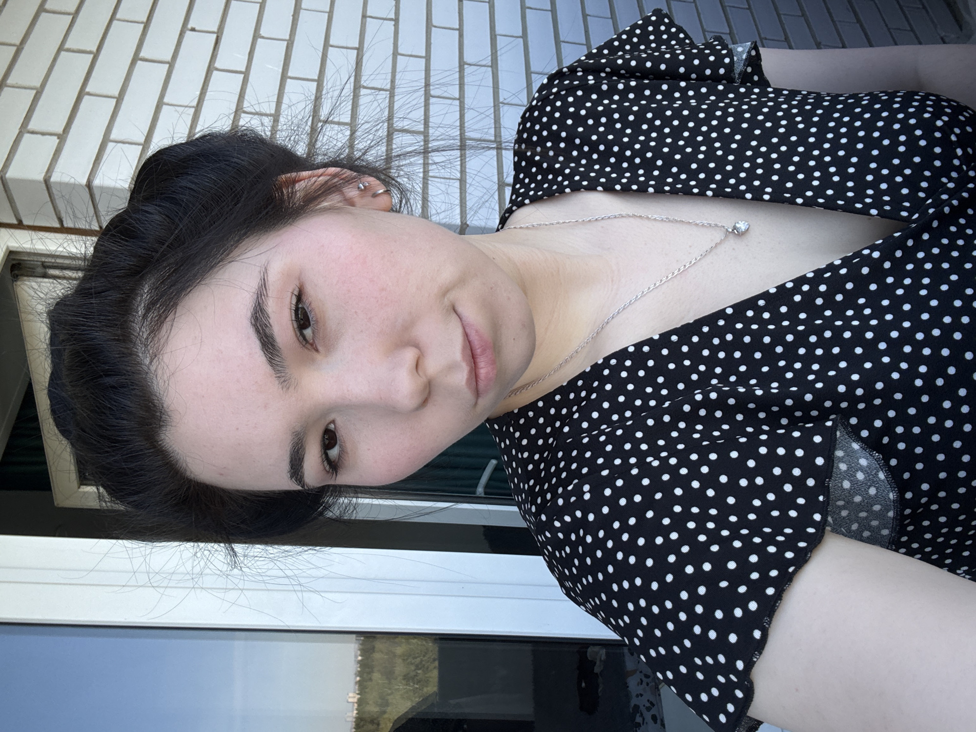 A young woman poses for a close-up selfie. She has dark hair styled in a bun, fair skin, and brown eyes. She wears a black dress with white polka dots and a silver necklace with a pendant. A brick wall is visible in the background, along with what appears to be a window reflecting a cityscape. The picture provides no indication of the city of Izhevsk, Russia.
