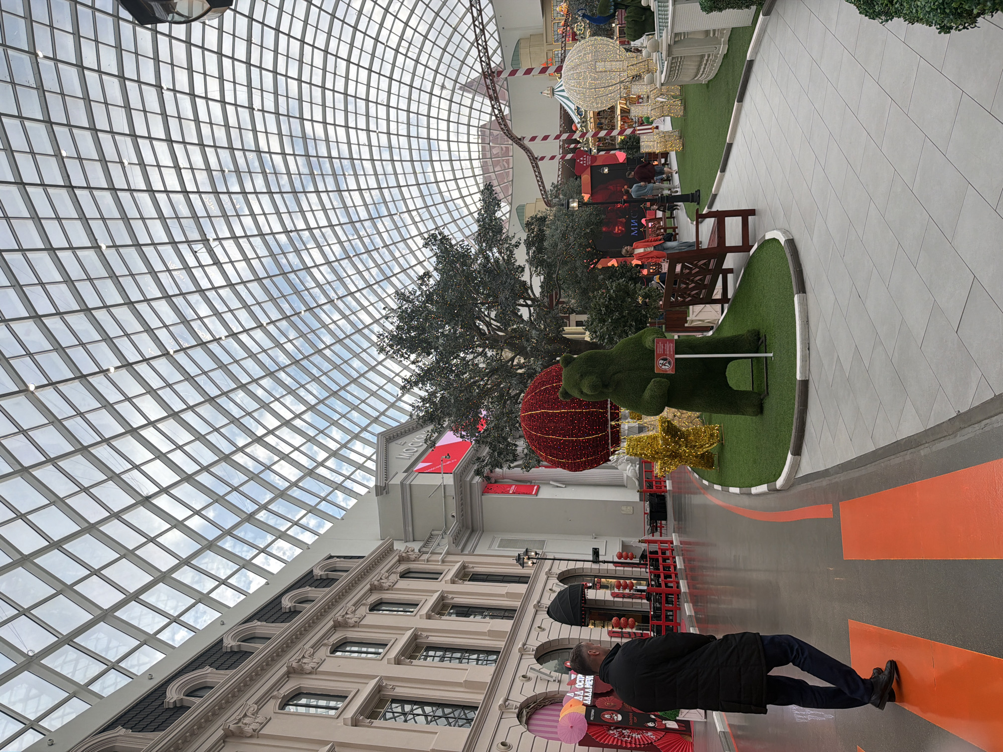 This image depicts the interior of a large, modern building with a distinctive geodesic dome glass roof. The scene is likely a shopping mall or entertainment complex, indicated by the presence of various decorative elements and retail-like displays.
The subjects include several people walking and observing the surroundings. One person in the foreground is facing away from the camera, wearing a black jacket and dark pants. Other individuals are visible in the middle ground, interacting near some of the displays.
Objects in the scene include large, festive decorations such as a giant illuminated sphere and a star-shaped light structure, suggesting a holiday or special event. There is also artificial grass landscaping, benches, and a roller coaster track visible in the background, hinting at an amusement or entertainment aspect to the venue. A large, stylized tree is also a prominent feature.
The setting is an indoor environment with a bright, natural light filtering through the glass dome. The architecture features classical-inspired facade details on the buildings within the complex. The floor is paved with light-colored tiles and has an orange pathway painted on it.
There are several pieces of visible text. A sign on the building reads "МОСКО". Another sign on the green topiary figure appears to be a warning or informational sign with a symbol. Some decorative banners or signs with what appears to be Asian-inspired imagery are also visible near the bottom of the frame.
The overall impression is one of a vibrant, possibly festive, indoor public space with a mix of shopping, entertainment, and decorative elements. The time of day appears to be daytime, with the sky visible through the dome indicating clear or partly cloudy weather. There are no strong emotional cues conveyed by the people depicted.