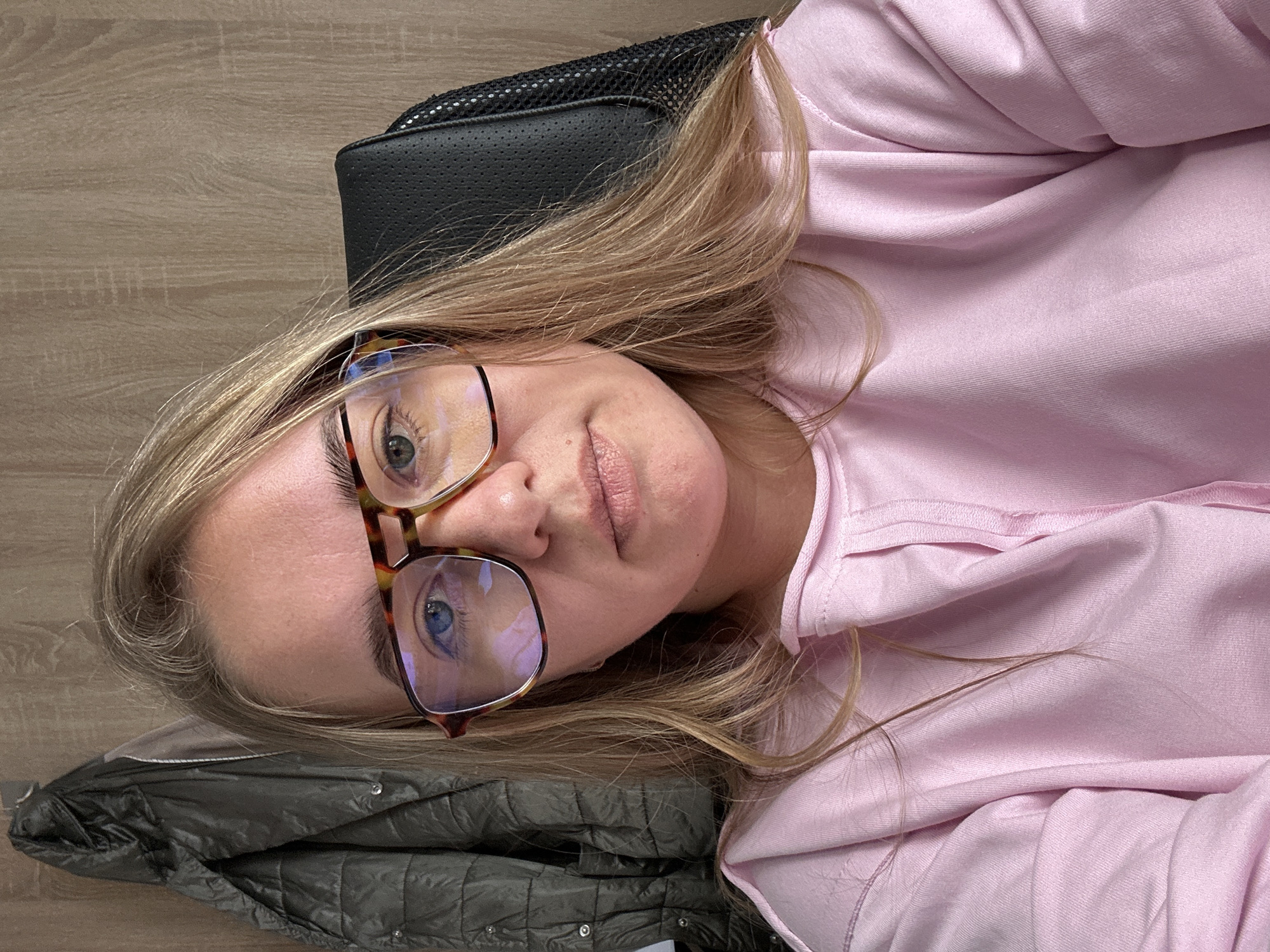 A person with light skin, blonde hair, and blue eyes is visible in a selfie-style photograph. The person is wearing eyeglasses with a tortoiseshell pattern frame and a light pink hooded sweatshirt. They are reclining against a black, perforated material that appears to be the headrest of a chair. A folded gray jacket with a quilted pattern is beneath the person's head. The background consists of a wooden-patterned surface, which may be a table or floor. No indication shows Ural'skoe, Russia.