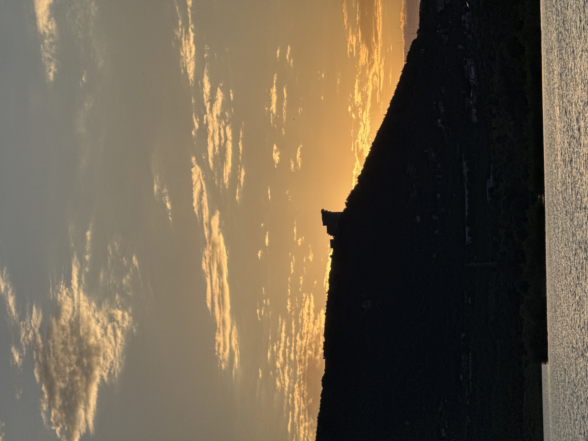 A silhouetted medieval fortress stands atop a dark, grassy hill overlooking a body of water. The sky above is painted with the warm hues of sunset, with scattered clouds catching the golden light. The fortress, perched on the peak of the hill, is a prominent structure, its distinct shape outlined against the vibrant sky. The hill itself is covered in dark vegetation, and the lower slopes show evidence of buildings and structures, suggesting a town or settlement. The water in the foreground reflects the colors of the sky, creating a shimmering surface. The overall scene evokes a sense of historical grandeur and natural beauty. This is a view of Shumen Fortress in Shumen, Bulgaria.