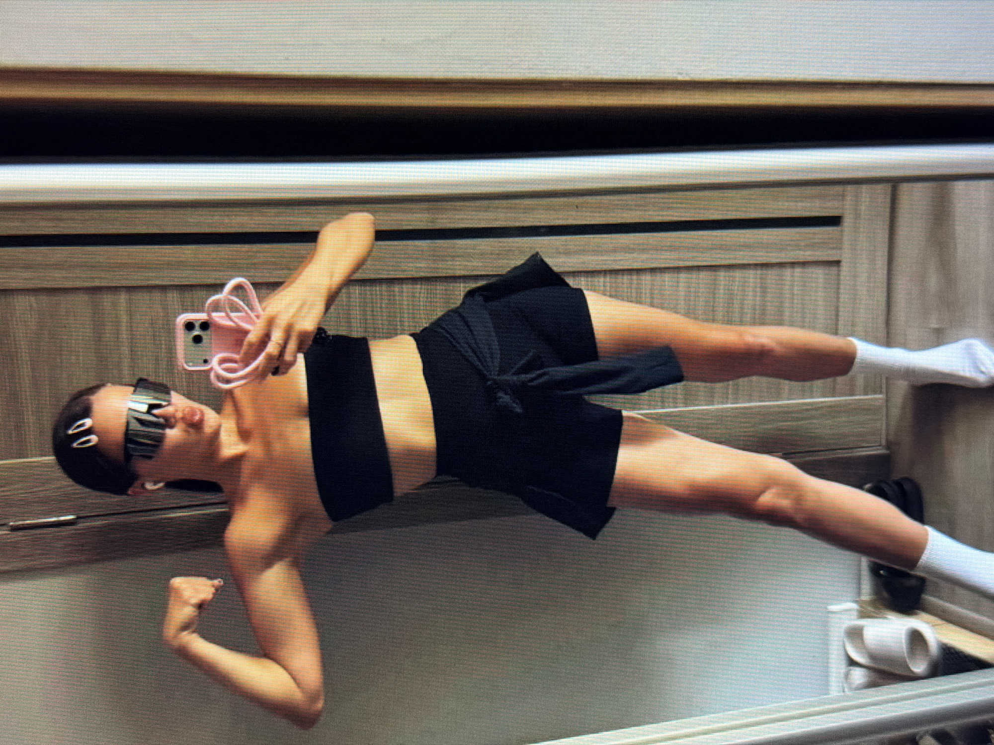 This media file shows a woman taking a mirror selfie in what appears to be a bedroom. She is wearing a black sports bra, black shorts, and white socks. She has on futuristic-style sunglasses and her hair is pulled back with two white clips. She is holding a pink phone with a heart-shaped strap in her right hand and is flexing her left arm. The background is a wooden wall with horizontal paneling. A white mug and what looks like a shoe are visible on a shelf or surface in the lower right corner. The lighting suggests it is daytime, and there are no clear indications of the weather or any specific emotions displayed beyond a confident pose. There is no visible text in the image. The location context provided is Sirius, Russia.