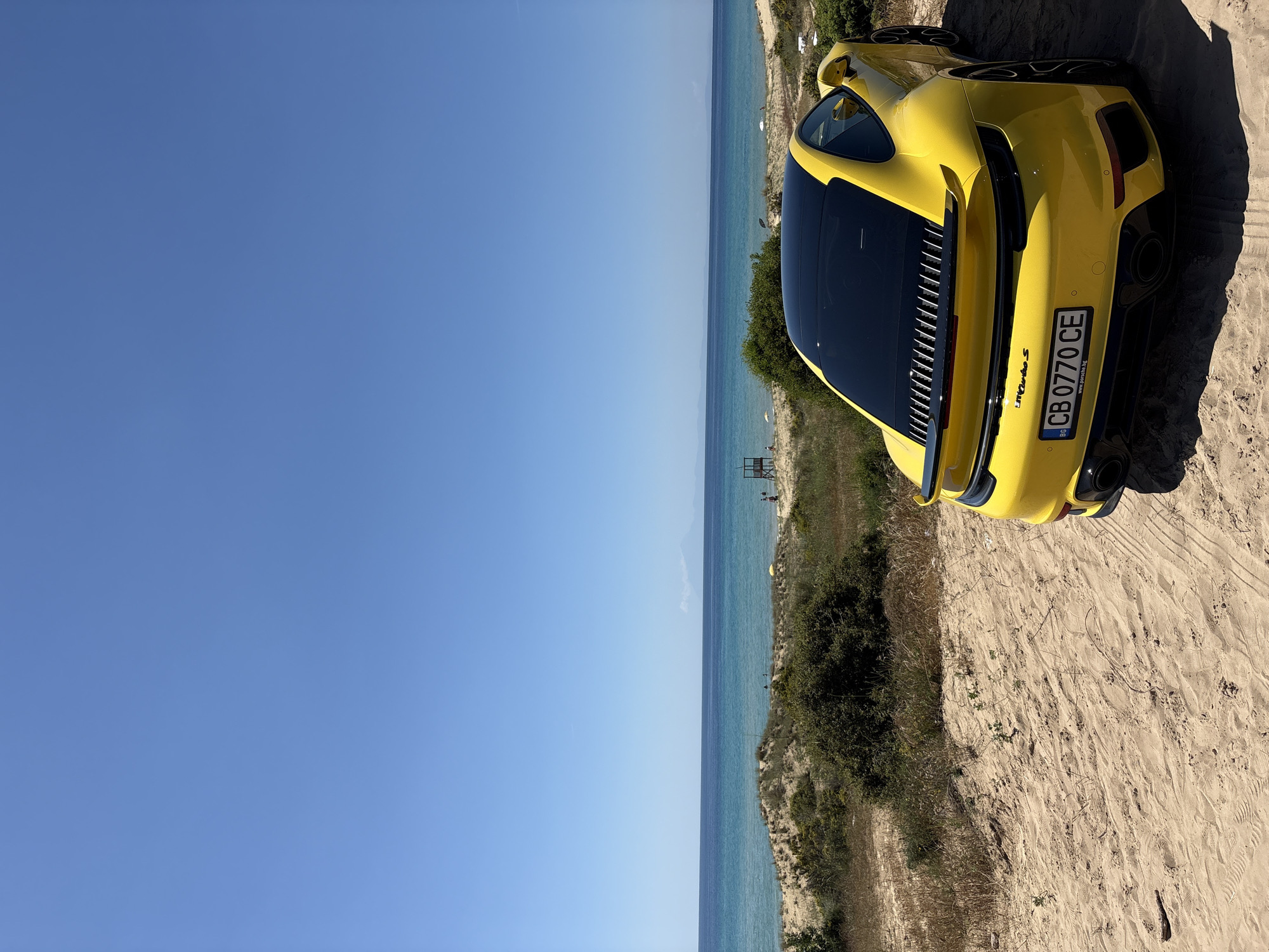 The image shows a yellow Porsche 911 Turbo S parked on a sandy area near a beach in Nea Peramos, Greece. The car is positioned at the edge of a dune, with vegetation nearby. In the background, the Aegean Sea is visible under a clear blue sky. A lifeguard tower can be seen along the beach. The car's license plate reads "CB 0770 CE".