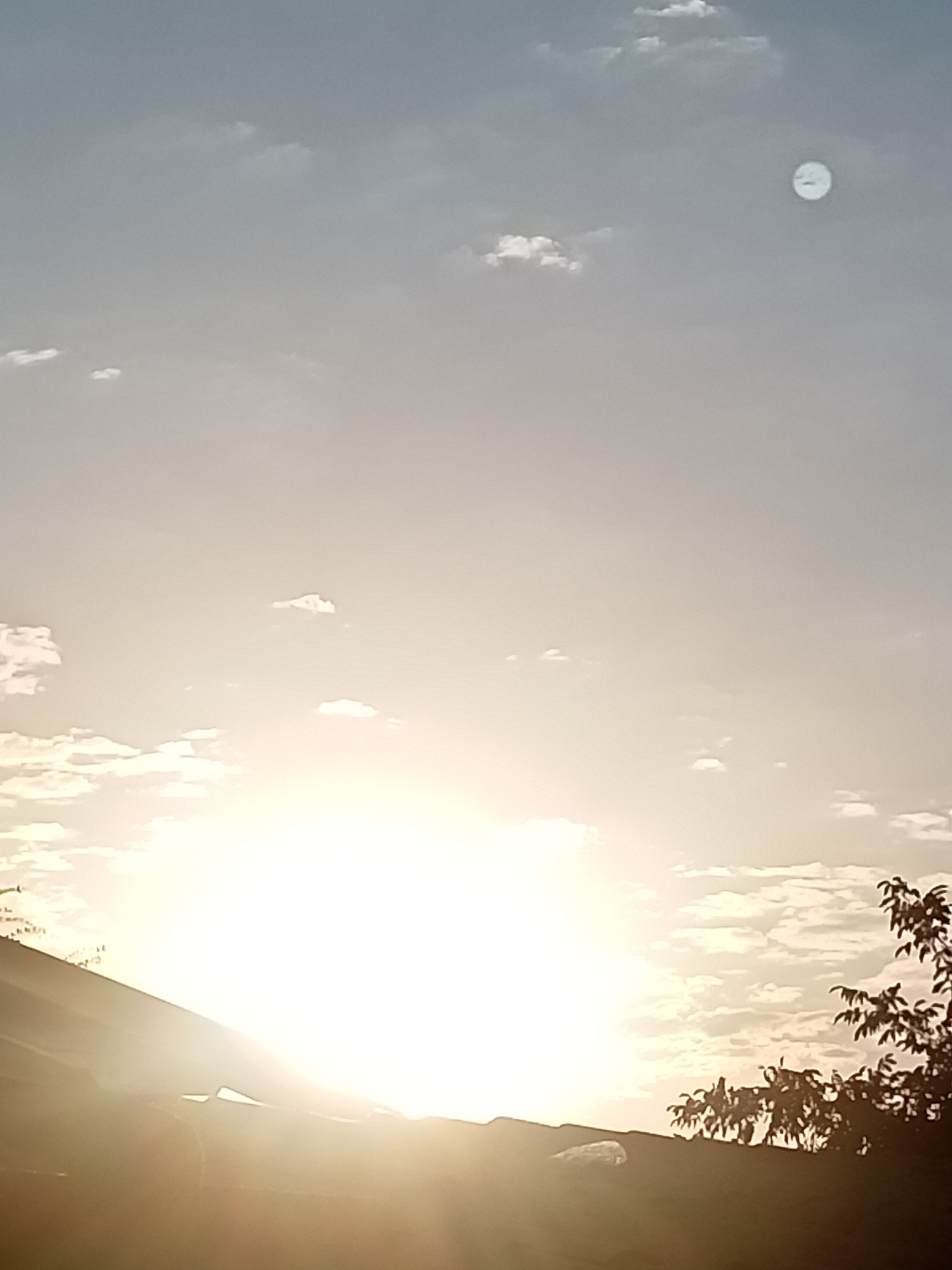 The image depicts a bright sky with scattered clouds. A large, bright celestial body, likely the sun, is positioned low in the sky, casting a strong glow that overexposes much of the lower portion of the image. The silhouette of a rooftop is visible in the foreground, partially obscuring the bright light. To the right, the dark silhouette of tree branches with leaves extends into the frame. A small, indistinct circular object is visible in the upper right portion of the sky, appearing lighter than the surrounding sky. The overall lighting suggests it is either sunrise or sunset. The location of Sokuluk, Kyrgyzstan is not evident from the visual information in the image.
