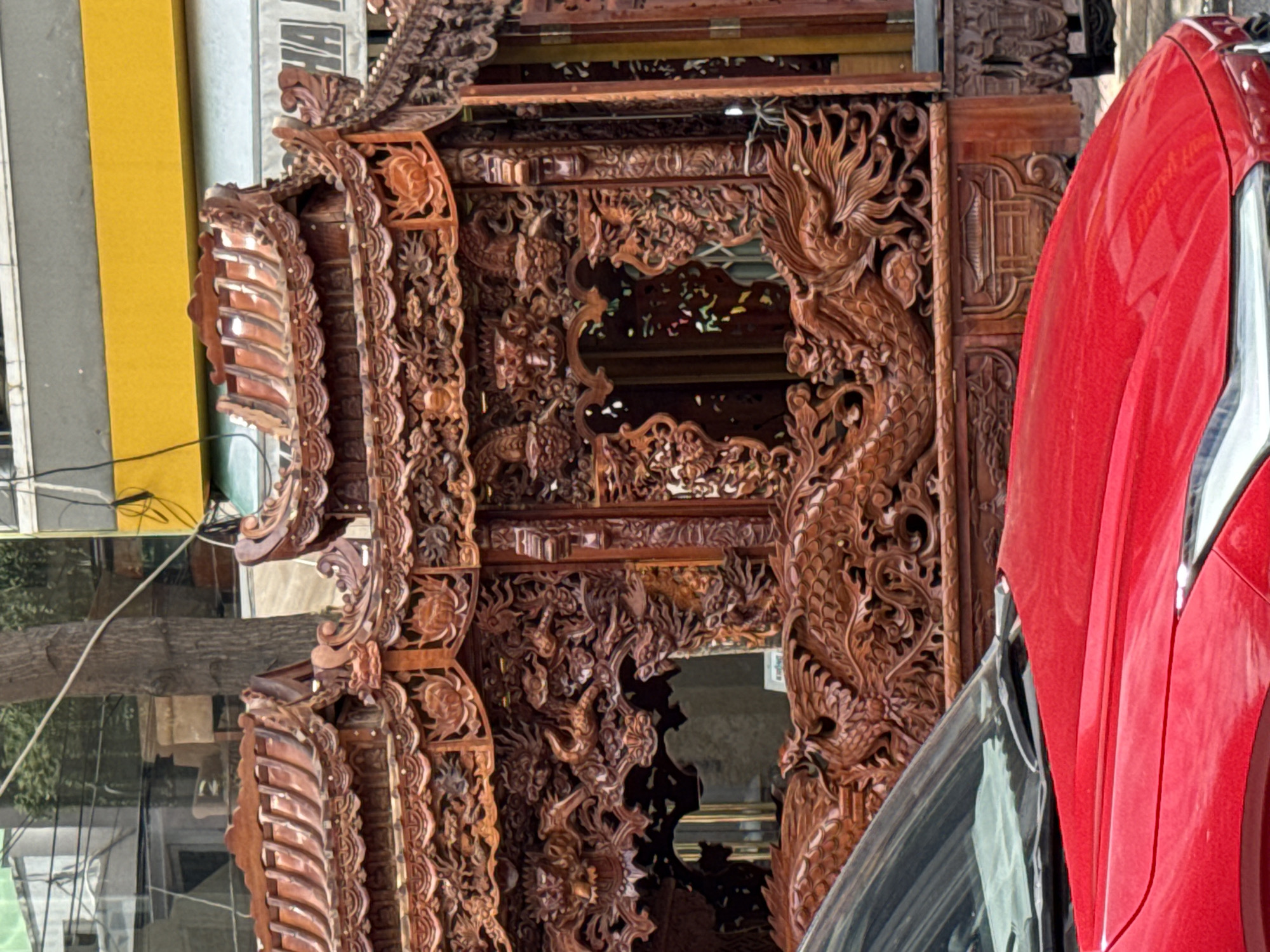 The image captures a street scene in Nha Trang, Vietnam, dominated by an intricately carved wooden structure, possibly a shrine or a float, featuring detailed dragon motifs and traditional architectural elements. The structure is made of dark reddish-brown wood and exhibits elaborate carvings of dragons, floral patterns, and what appear to be miniature pagodas. The roof has multiple tiers with curved eaves, typical of East Asian architecture. In the foreground, the hood and windshield of a red car are visible, partially obscuring the lower portion of the wooden structure. The background reveals a typical urban environment with buildings, trees, and wires, suggesting the scene is outdoors in a city. The lighting suggests it is daytime.