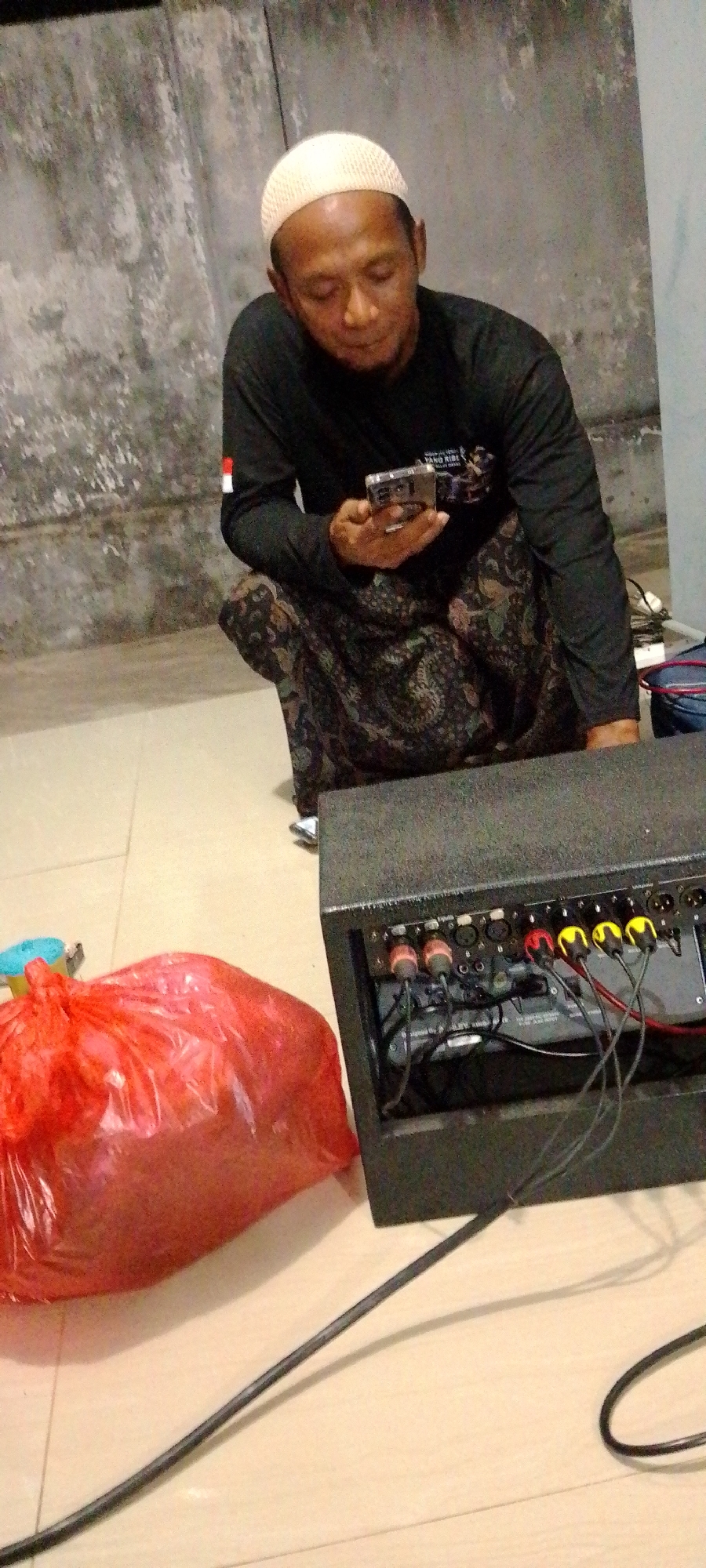 An adult male wearing a white peci cap and a black long-sleeved shirt with a patterned sarong is crouched on a tiled floor. He is holding a smartphone in his hands and appears to be looking at it. In the foreground, to the left, is a large red plastic bag. To the right is a black audio speaker with various cables and connectors visible on its back panel. The background consists of a textured wall. The scene is indoors, with no windows visible.