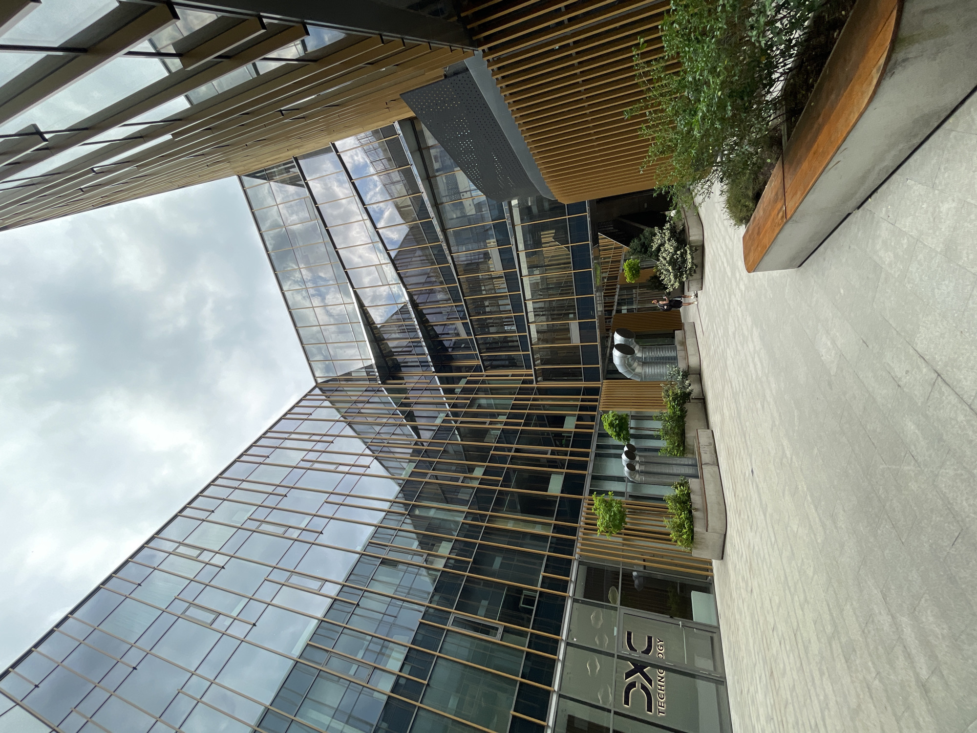 The image shows an exterior view of a modern building in Sofia, Bulgaria, likely an office complex. The building has a facade dominated by glass panels with a grid-like framework. The glass reflects the sky, suggesting cloudy weather. Part of the building has a wooden slat-like structure overhead. On the ground level, there's a walkway paved with light-colored tiles, and there are sections with plants and small trees. There is a sign indicating "DXC Technology" on the ground floor. Two people are walking in the background. The viewpoint is from below, looking upwards at an angle. There are vent-like structures in the background as well.