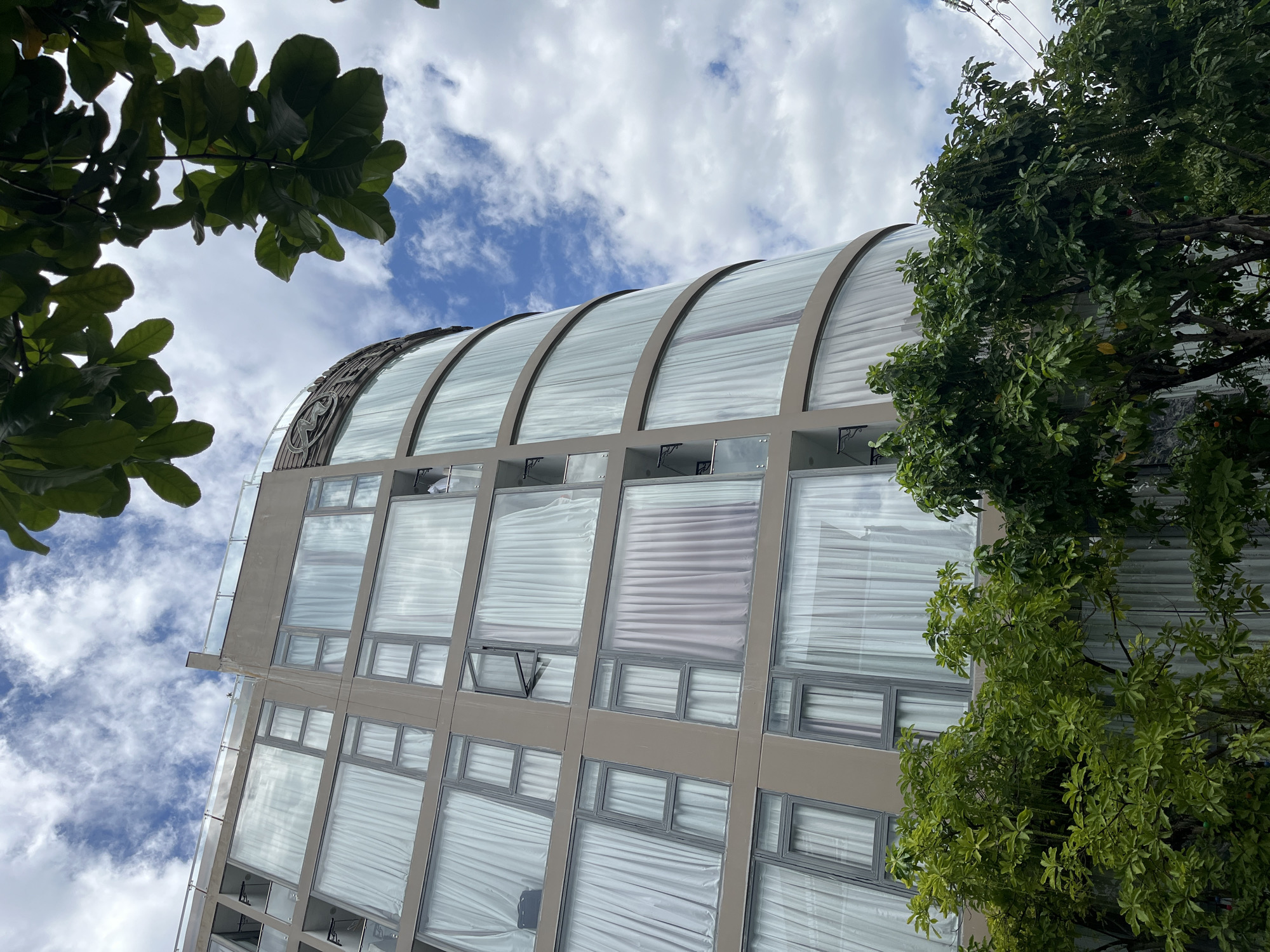 The image captures a modern, multi-story building from an upward perspective, set against a bright, partly cloudy blue sky. Lush green foliage from trees frames the structure, with branches extending from the left foreground and along the right side of the building.
The building's most prominent feature is its distinctive curved roof, constructed from translucent glass panels and supported by a light-colored framework. A stylized emblem, resembling an "M" or "W," is visible as a decorative element on the curved roof. Below this, the building's facade is dominated by numerous rectangular windows arranged in a grid. Most of these windows are covered by white or light-colored curtains, giving a uniform appearance. One smaller window on a lower floor is slightly open. The building's exterior walls between the window sections are a light beige or gray. Some decorative brackets or structural supports are visible beneath the upper windows. Faint electrical wires are discernible against the sky in the upper right corner.
The scene depicts a clear, bright daytime with natural lighting. There are no individuals visible, and no particular event or activity is taking place. The modern architecture combined with the prominent tropical greenery is consistent with the provided location context of Đà Nẵng, Vietnam.