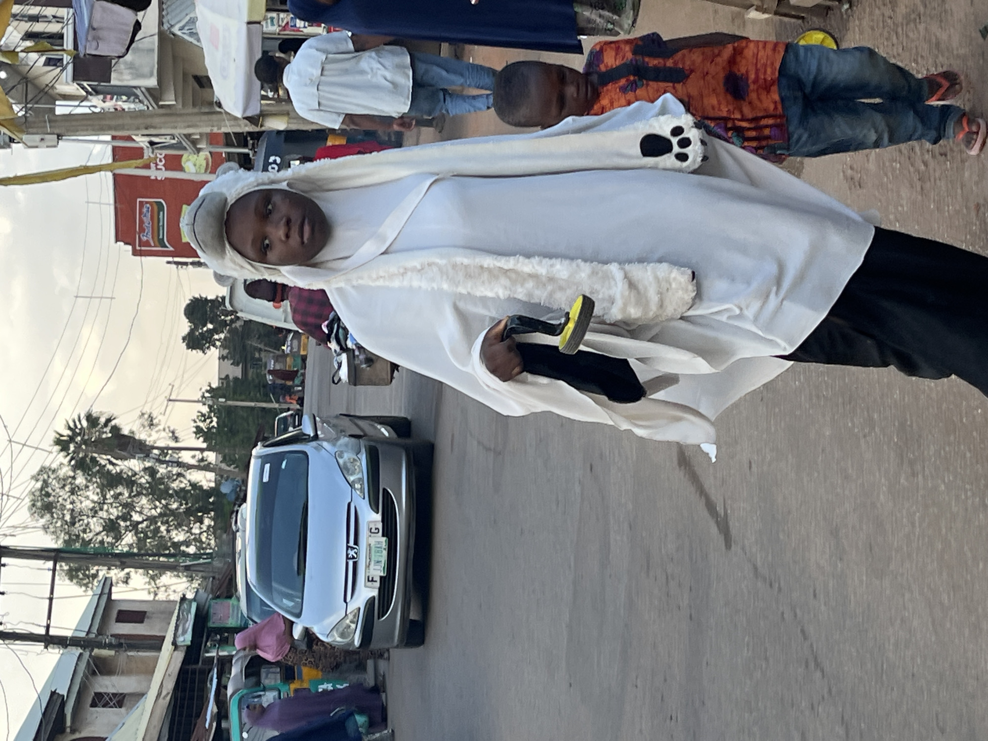 In Jos, Nigeria, on a street, a woman wearing a white hijab with a polar bear-like hood is walking. She has a fluffy white scarf draped around her neck, which is adorned with paw prints. In her right hand, she carries a black item with a yellow circular end. Beside her, a young boy in an orange and black patterned shirt and blue jeans walks, wearing pink flip-flops. The street is paved, and buildings line the right side of the image. A silver Peugeot car is visible in the background. Overhead power lines crisscross the sky. The scene appears to be a bustling market area.