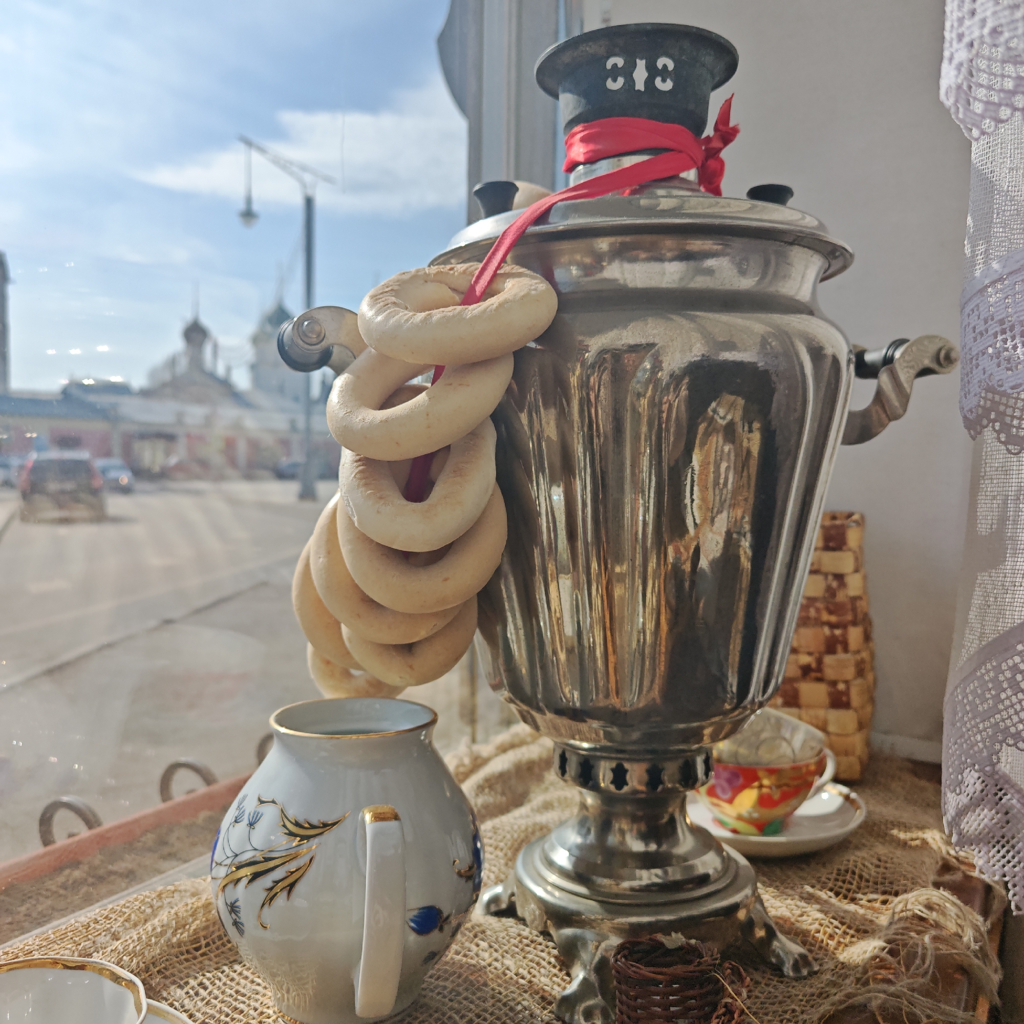 A medium indoor shot shows a samovar with a string of bagels hanging from it, displayed on a counter in front of a window overlooking a street scene in Rostov, Russia.
The samovar, made of shiny metal, is prominently positioned in the center of the frame. A red ribbon is tied around the top, and a string of bagels is draped over it. To the left of the samovar is a porcelain pitcher with gold trim and blue floral designs. A tea cup and saucer are to the right of the samovar. The samovar, pitcher, and tea cup rest on a coarse, burlap-like surface.
The background features a window with a view of a street in Rostov, Russia. Buildings with distinctive Russian architecture are visible across the street. Cars are moving on the street, and streetlights and overhead wires are visible.
The lighting is soft and natural, coming from the window in the background. The overall impression is that of a traditional Russian tea setting displayed as a window display.