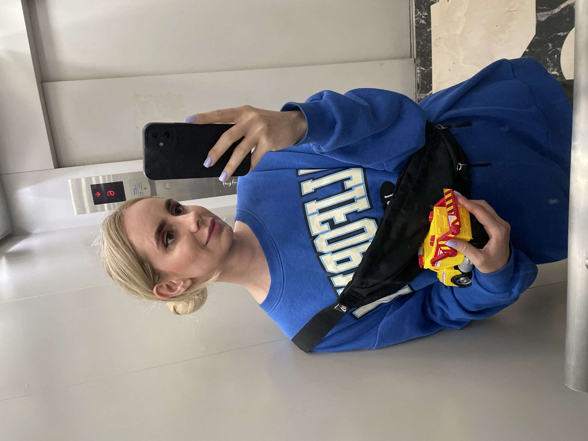 A woman is taking a selfie in an elevator. She has blonde hair pulled back in a bun and is wearing a blue sweatshirt with a black fanny pack. She is holding a toy fire truck in one hand. The elevator has white walls and a digital display showing the floor number. The exact location in Vladivostok, Russia is impossible to determine from the image.