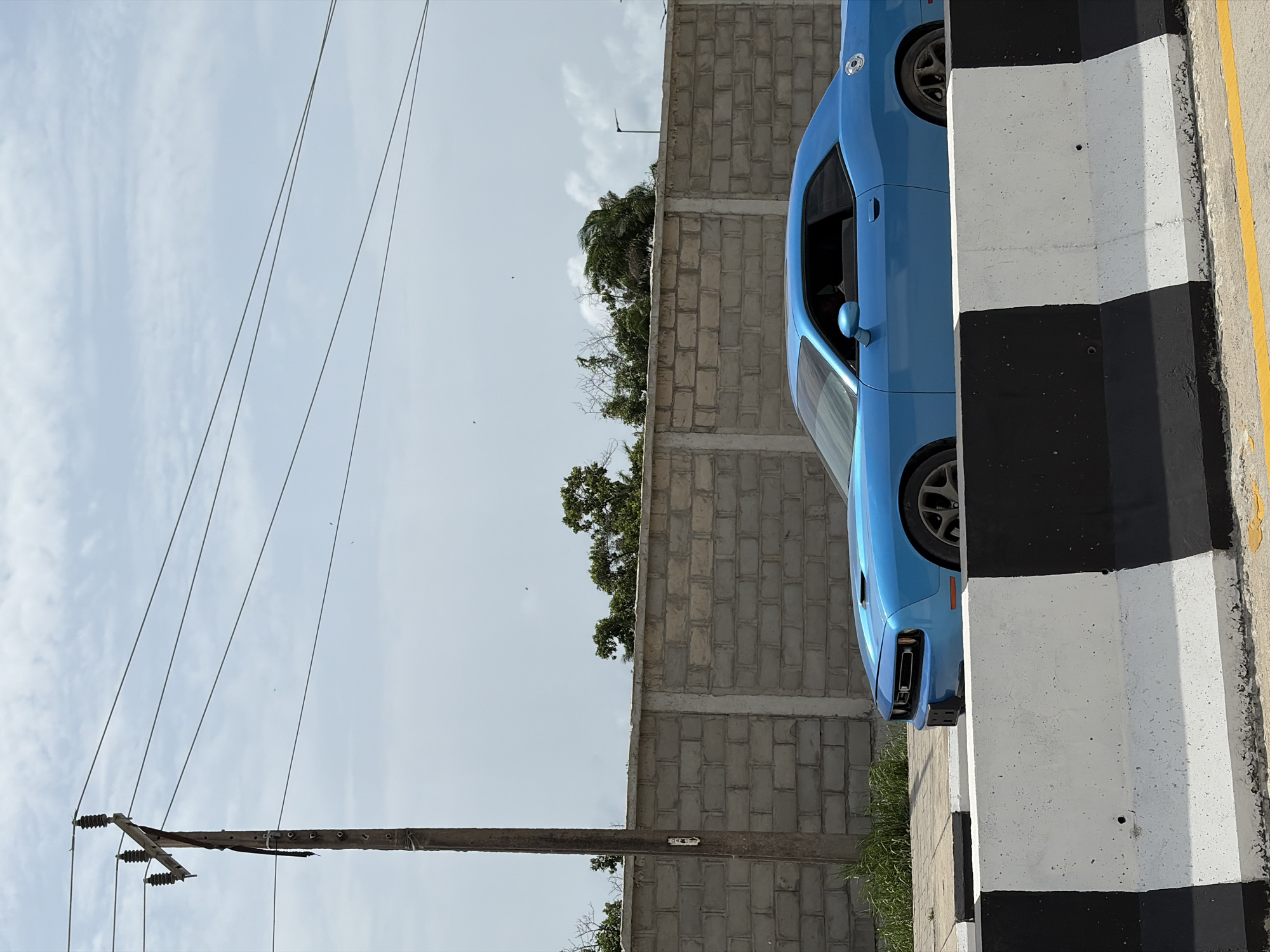 The image, rotated 90 degrees, depicts a blue car parked near a black and white striped barrier. The car is positioned next to a concrete block wall. In the background, there's vegetation, including trees. Electrical power lines are visible in the left background, supported by a utility pole. The location is potentially Igboefon, Nigeria.