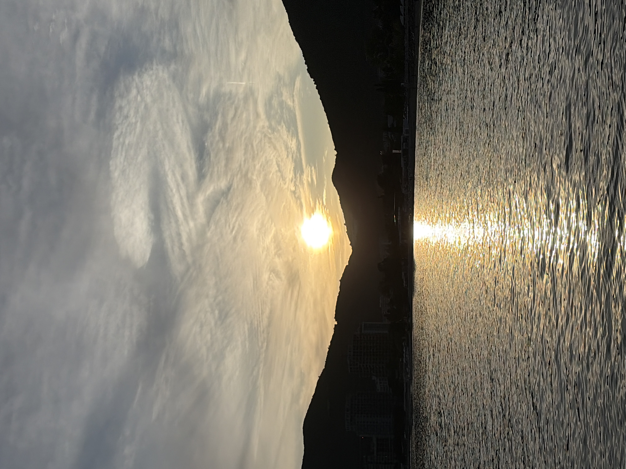 The image captures a sunset over the Adriatic Sea in Budva, Montenegro. The sun is setting behind a mountain range, casting a golden glow on the water. The water is rippled, reflecting the sunlight in a shimmering pattern. In the foreground, there is a glimpse of the city's coastline with buildings visible along the shore. The sky is filled with clouds, creating a dramatic and colorful backdrop. The image is rotated 90 degrees counterclockwise.