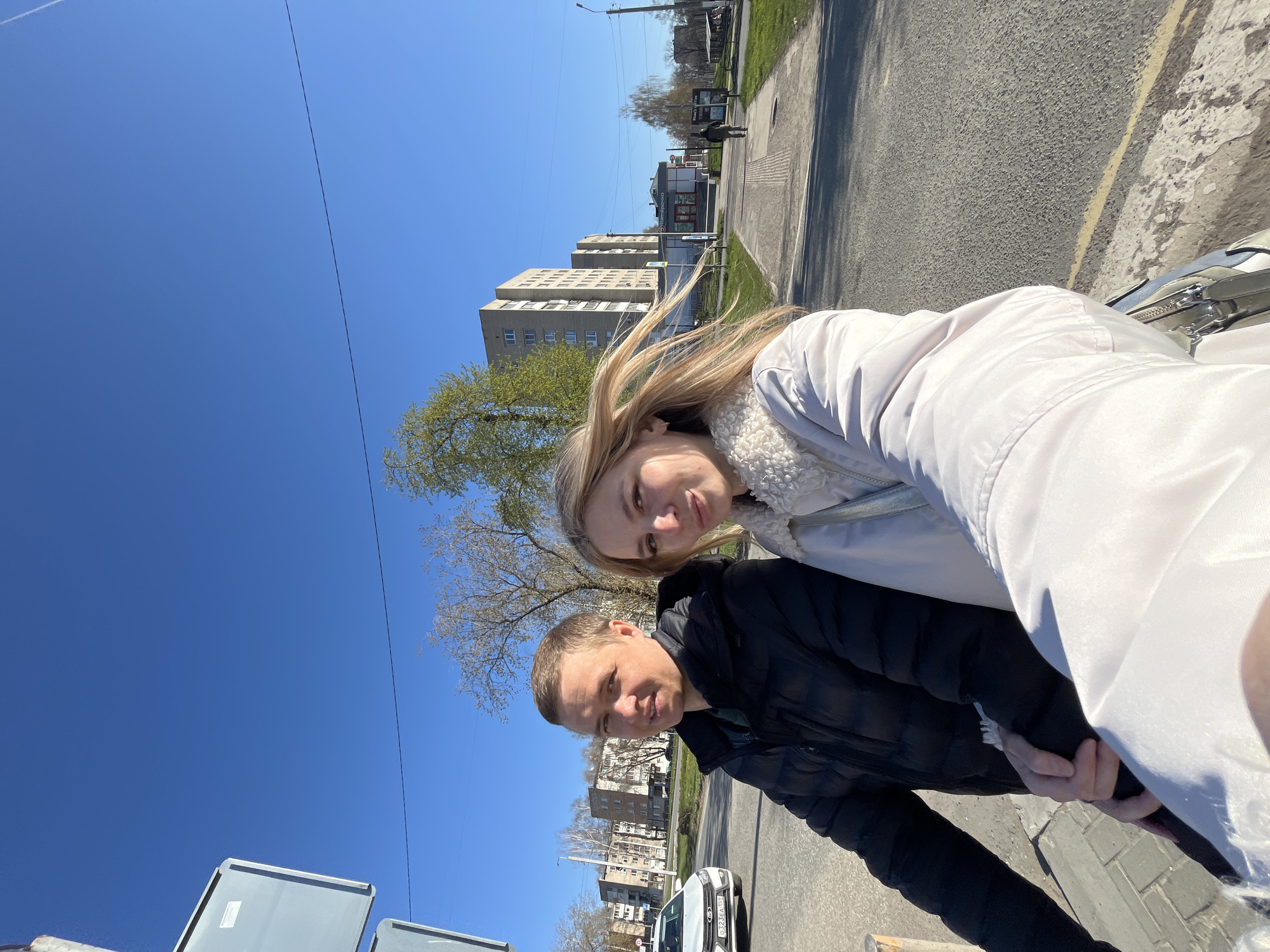 A couple poses for a selfie along a street in Chaikovsky, Russia. The woman is on the right, wearing a white jacket, and the man to her left is in a black jacket. They are standing on a sidewalk. Behind them is a street with a few cars and pedestrians. Buildings are visible in the background, and the sky is blue with a few trees.