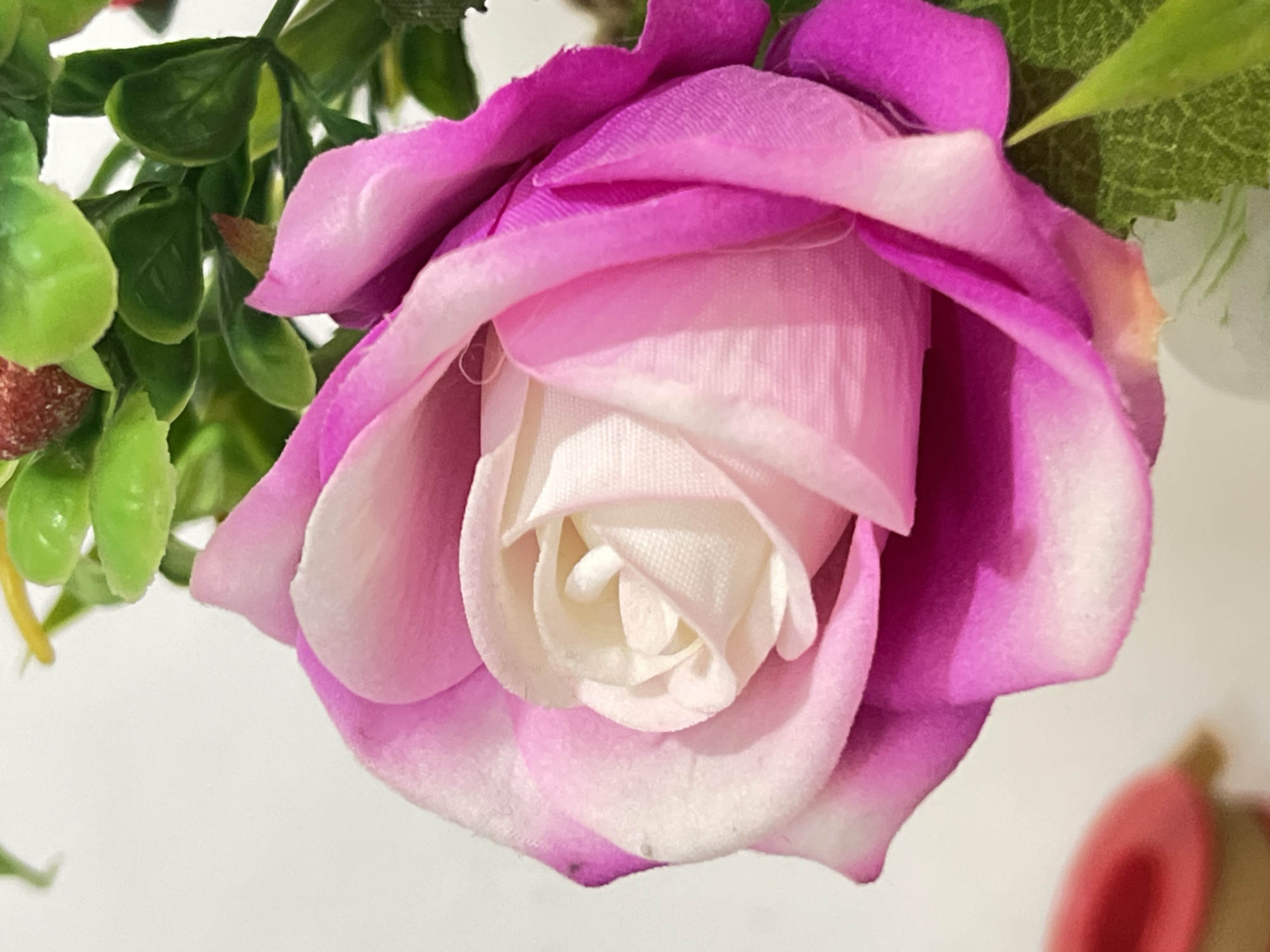 This is a close-up, vertical shot of a pink and white rose. The rose is artificial, with fabric petals that are soft and velvety in texture. The inner petals are white, tightly wound in the center, and transition to a soft pink, then to a vibrant fuchsia on the outer petals. The rose is surrounded by green artificial leaves and a hint of red on a berry-like object is visible in the upper right portion of the image. The background is a plain, off-white color. The scene appears to be indoors, possibly in a home or a shop in Denpasar, Indonesia, where artificial flowers are commonly sold or used for decoration. There are no people present in the image.