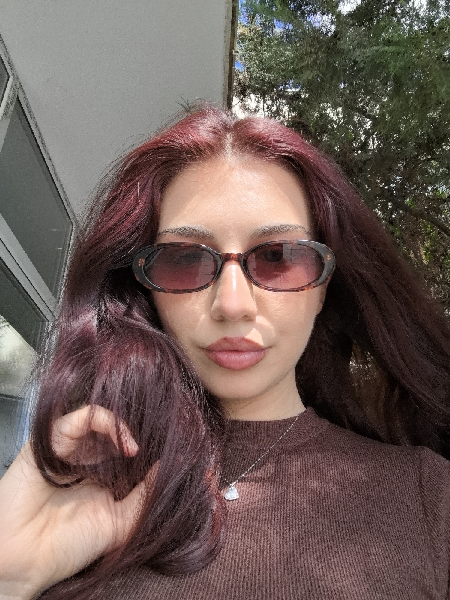 A woman with long, burgundy hair and tortoise-shell sunglasses is the primary subject of the close-up photograph. She is wearing a brown, ribbed, high-necked shirt and a silver necklace with a heart-shaped pendant. Her right hand is visible, slightly obscuring part of her hair and shirt. The background includes a white building with windows and some green foliage, suggesting an outdoor setting. The city and specific location are unknown.