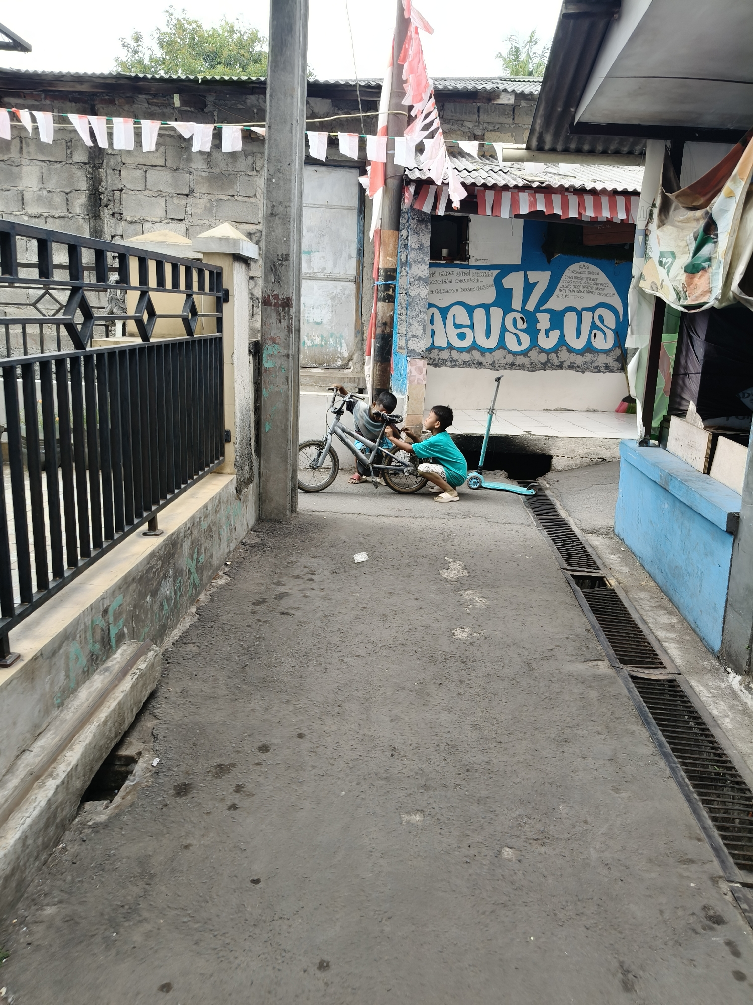 The image depicts an alleyway in South Tangerang, Indonesia, likely near a residential area. Two young boys are present. One boy, wearing a teal shirt and white shorts, is crouched beside a small bicycle, appearing to interact with it. Another boy, whose clothing is mostly obscured by the bicycle, is also near it. A light blue kick scooter is parked to the right of the bicycle.
The alley is paved with asphalt and has a concrete drainage channel running along its right side. A black metal fence borders the left side of the alley, behind which is a low concrete wall and some greenery. The background shows buildings with corrugated metal roofs and brick walls. Indonesian flags, red and white, are strung across the scene, suggesting a celebration or event, possibly Indonesian Independence Day on August 17th, as indicated by the mural on the building in the background that reads "17 AGUSTUS". The overall environment appears to be a modest urban setting.