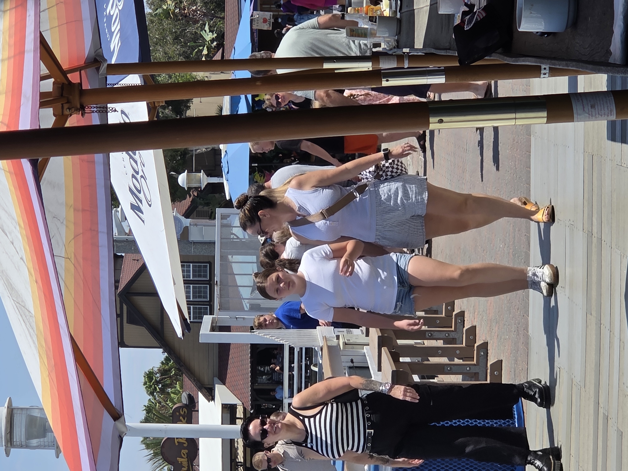 This image captures a sunny outdoor scene at the Pike Outlets in Long Beach, California. The foreground is dominated by a wooden boardwalk and part of a table with various items. Several people are walking, with two young women in the center being the most prominent. The woman on the left is wearing a black and white striped tank top, black pants, and black boots. The girl next to her is in a white t-shirt and denim shorts. To the right, another woman is seen wearing a light blue tank top and striped shorts. In the background, there are structures with awnings, one featuring colorful stripes and another with a "Modelo Especial" logo. Palm trees and other foliage are visible, indicating a warm climate. People are seen in various states of activity, suggesting a bustling commercial area or public gathering space.