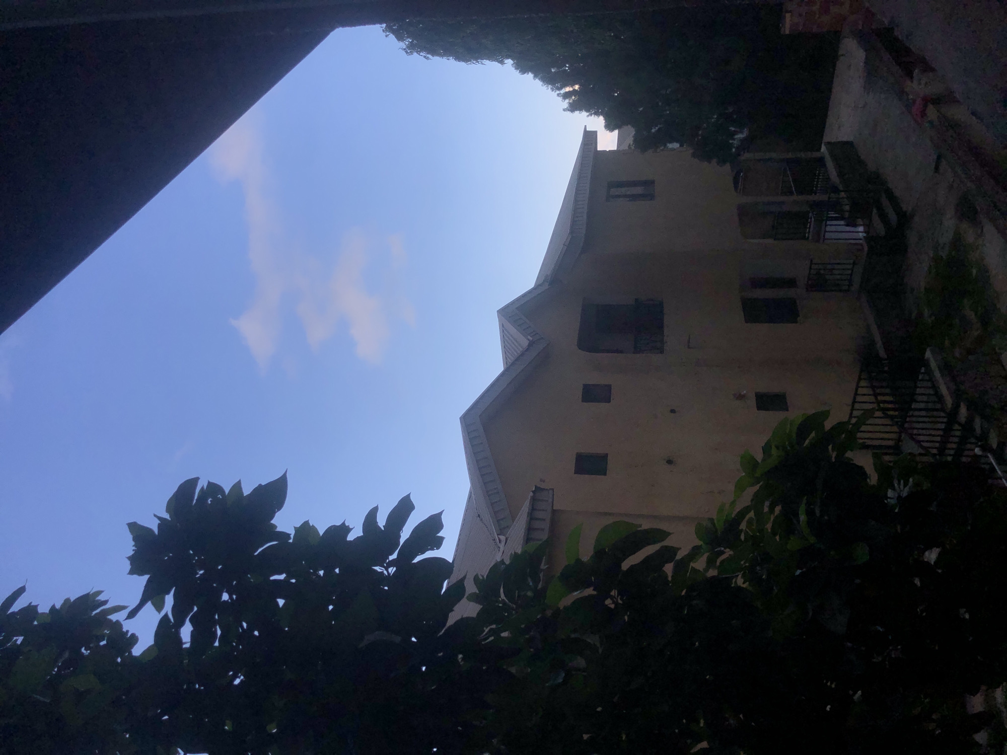 This is an outdoor, eye-level, medium shot of a two-story house in Barakin Lerawi, Nigeria, under a cloudy blue sky. The house is painted a light tan color and has a metallic gray roof. To the left of the house, a large tree with dark green leaves partially obscures the view. To the right, a tall, dark green tree is visible, and a dark brown overhang extends from the top right corner of the frame. The foreground is dimly lit, showing concrete stairs with black railings leading up to a porch area. Several small, dark rectangular windows are visible on the house, and a balcony with a railing is on the second floor. The overall scene suggests a residential setting in a tropical or subtropical climate.