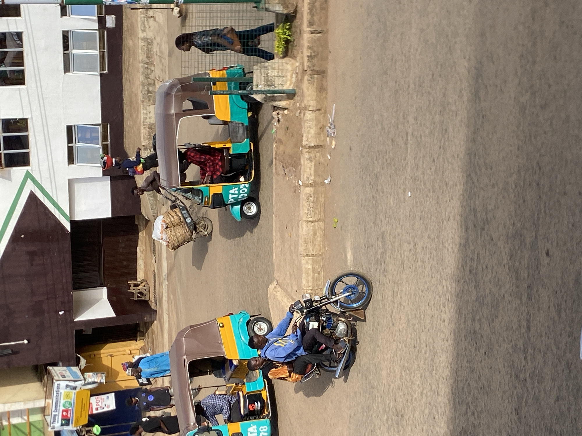 The image depicts a street scene in Bukuru, Nigeria. Two auto rickshaws, also known as "keke napep", are visible. One is green and yellow with a brown roof, containing a person in a red plaid shirt. The other, also green and yellow with a brown roof, carries at least two people. A motorcycle with two riders is also present. Other people are walking near the auto rickshaws. The buildings lining the street are a mix of white and brown, with windows visible in one of them. One building is also adorned with signage. The street appears paved, with some litter present. The photo seems to have been taken from an elevated vantage point.