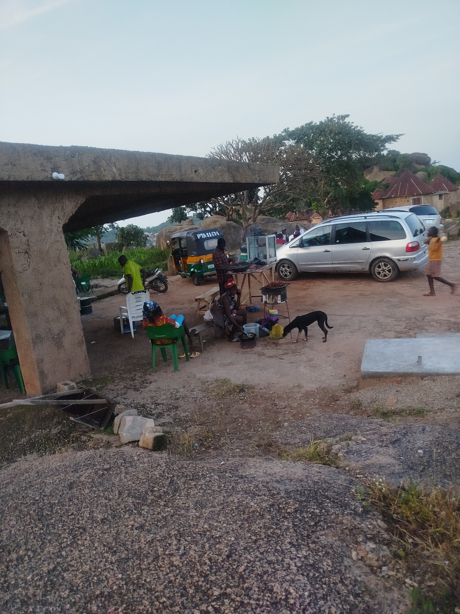 The image captures an outdoor scene at what appears to be a roadside market or resting spot on a rocky terrain in Jos, Nigeria. In the foreground, large granite boulders dominate the landscape, with sparse vegetation growing between them.
To the left, a concrete structure with a rough, unfinished texture extends from the bottom left corner, forming an overhang. Underneath this structure, several green plastic chairs are visible, one of which is occupied by a person with their back to the camera, wearing a colorful headscarf and a green top. Another person in a bright green shirt stands near a white plastic chair and a motorcycle.
In the middle ground, a yellowish-green three-wheeled auto-rickshaw, identified by the license plate "PTA-11674", is parked. Adjacent to it, a silver minivan is also parked. Several people are gathered around a makeshift food stall. A person in a yellow t-shirt and brown shorts is walking away from the minivan towards the right side of the frame. A black dog is sniffing the ground near the food stall.
Further in the background, traditional round houses with conical roofs are nestled among trees and rocky outcrops, characteristic of the landscape around Jos. The sky is clear and pale blue, suggesting either early morning or late afternoon light. The overall impression is a modest, functional gathering point set against a rugged natural backdrop.