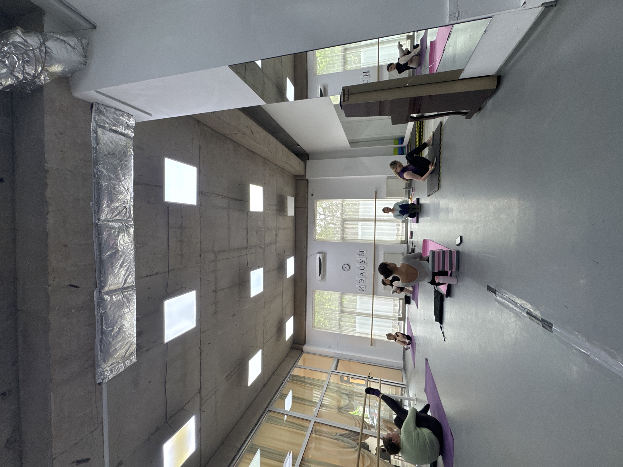A bright, well-lit room with a high ceiling is oriented vertically. Several people are engaged in stretching or yoga exercises on purple mats. They are aligned in a row towards a window along the right side of the image. The window is framed by orange-tinted glass panels. A ballet barre is also visible on the right. The floor appears to be a light gray color. The ceiling above is a mix of exposed concrete and drywall, with recessed square lights. A duct runs vertically along the left side of the image. In the distance, more individuals are participating in similar activities. A large mirror is visible on the right side of the image, reflecting the scene back into the room. The location is Unknown, Unknown.