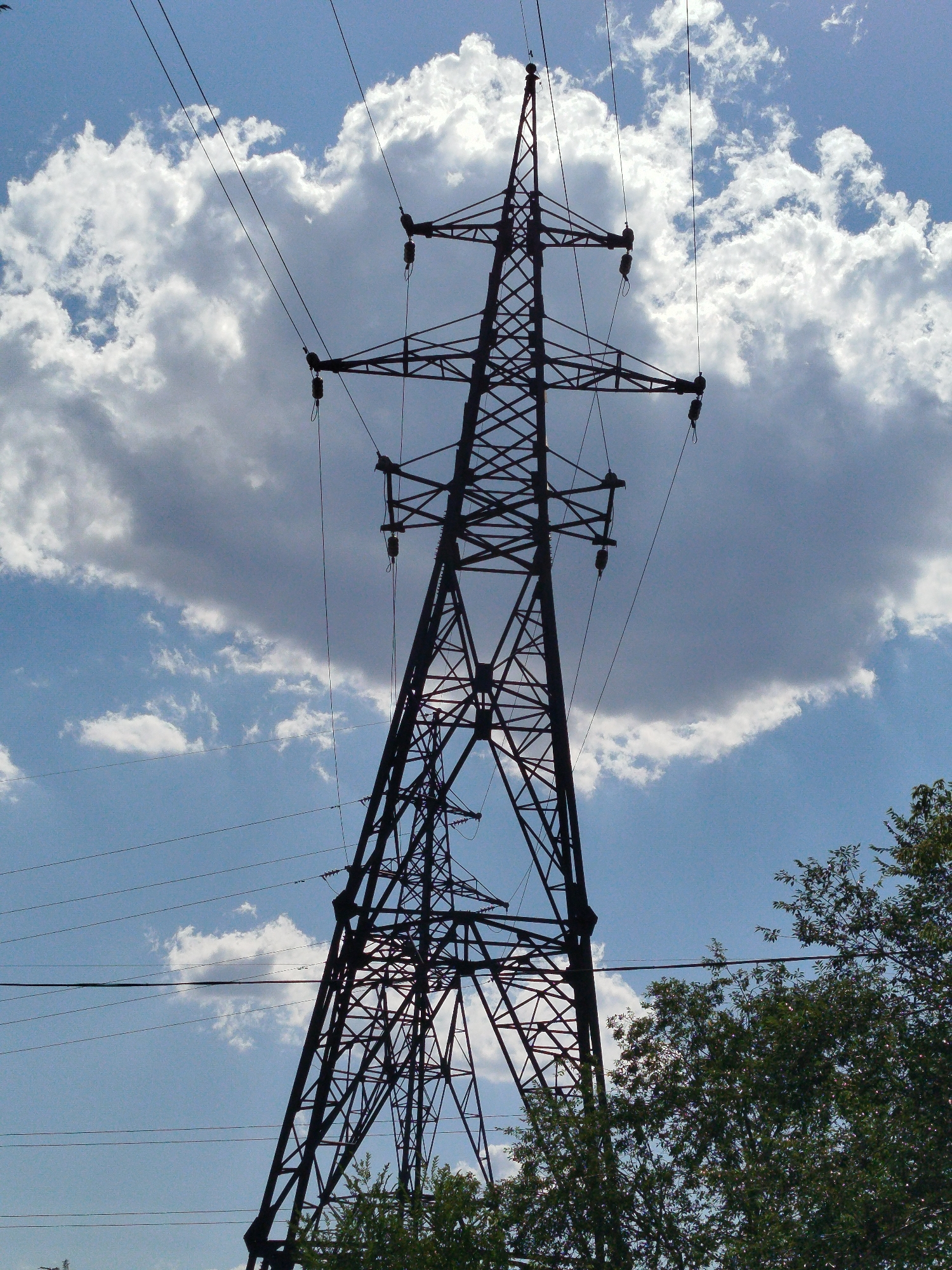 A low-angle, full shot captures a tall, dark metal power line tower silhouetted against a bright blue sky filled with fluffy white and gray clouds. The tower's complex lattice structure is visible, with multiple wires extending from it and disappearing into the distance. The bottom of the frame shows the tops of lush green trees. The scene appears to be outdoors, possibly in a semi-rural or industrial area of Qarağandı city administration, Kazakhstan. No people or animals are present in the image. The lighting suggests it is daytime, with the sun illuminating the clouds from behind.